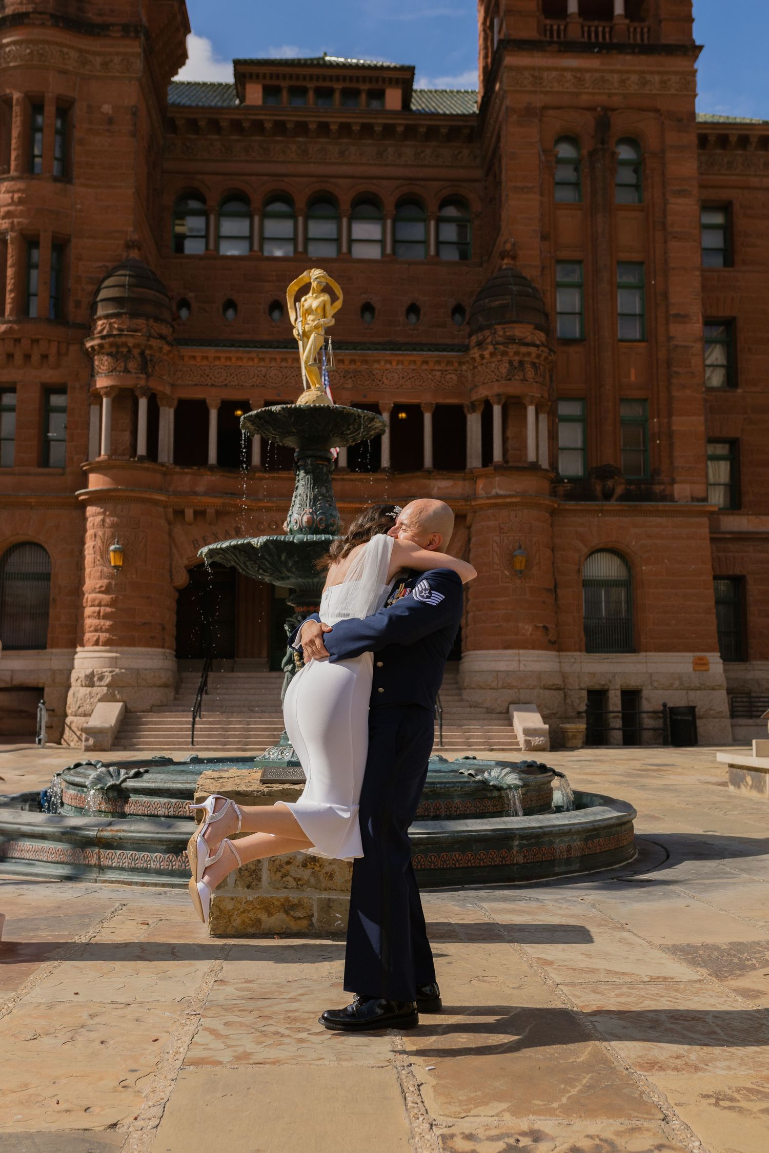 A romantic embrace by a fountain outside a historic red brick courthouse building.