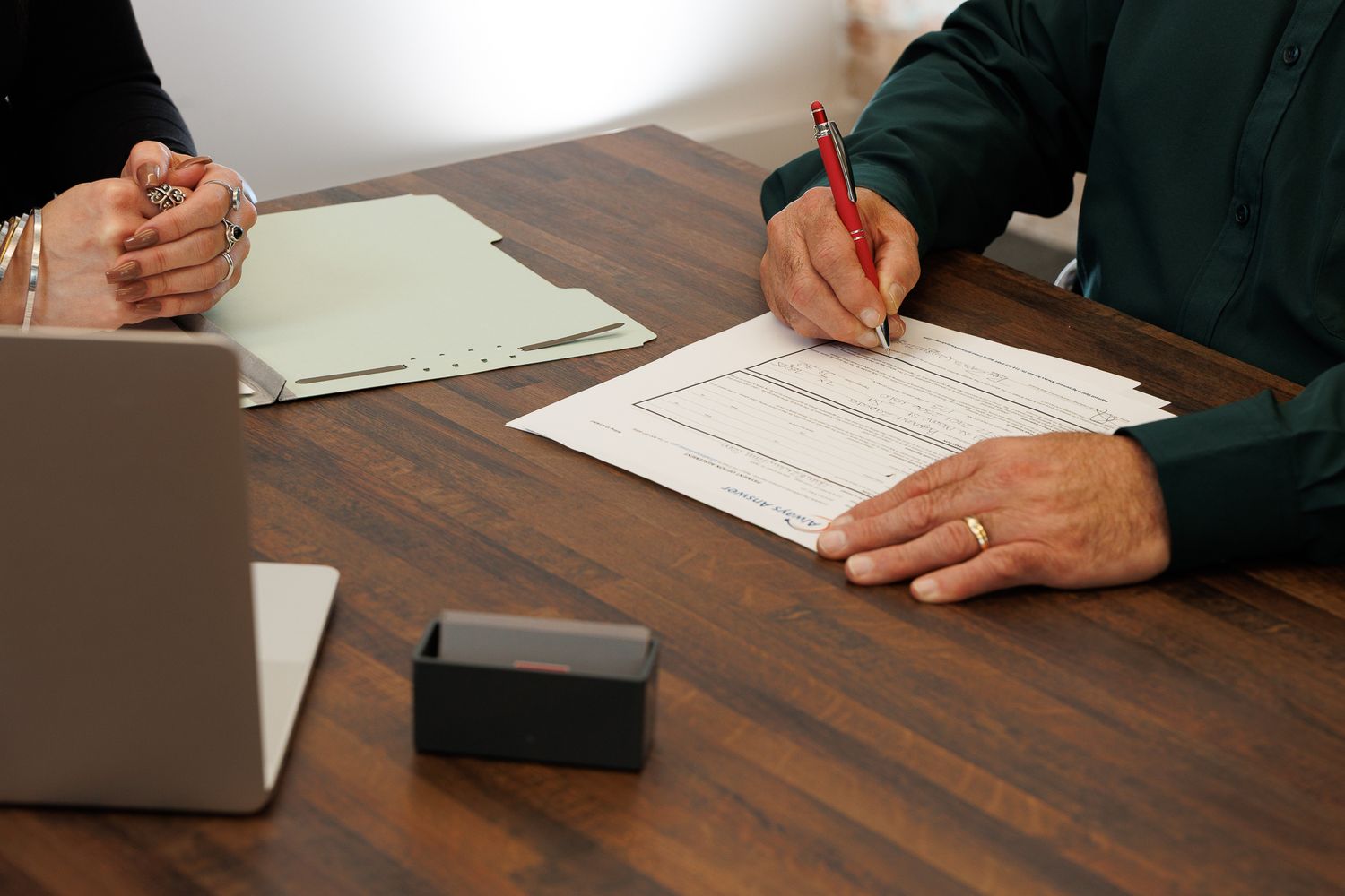 Two professionals signing documents at a wooden desk during a business meeting with a laptop nearby.