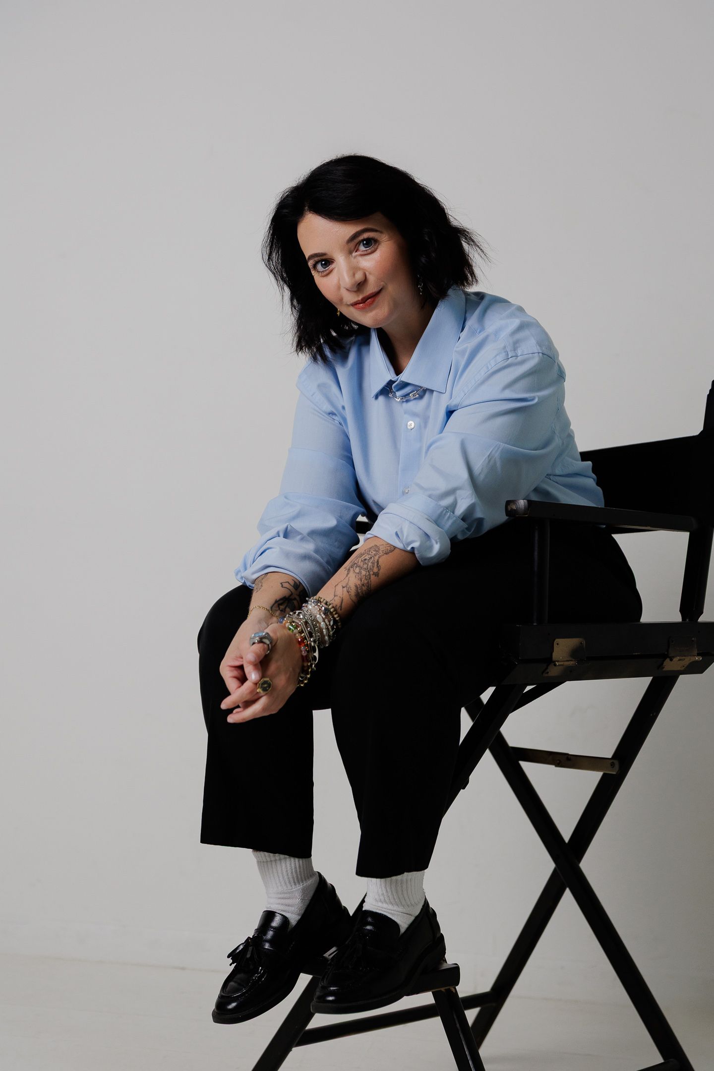 A person in a light blue button-down shirt and black pants sits casually on a director's chair against a gray backdrop.