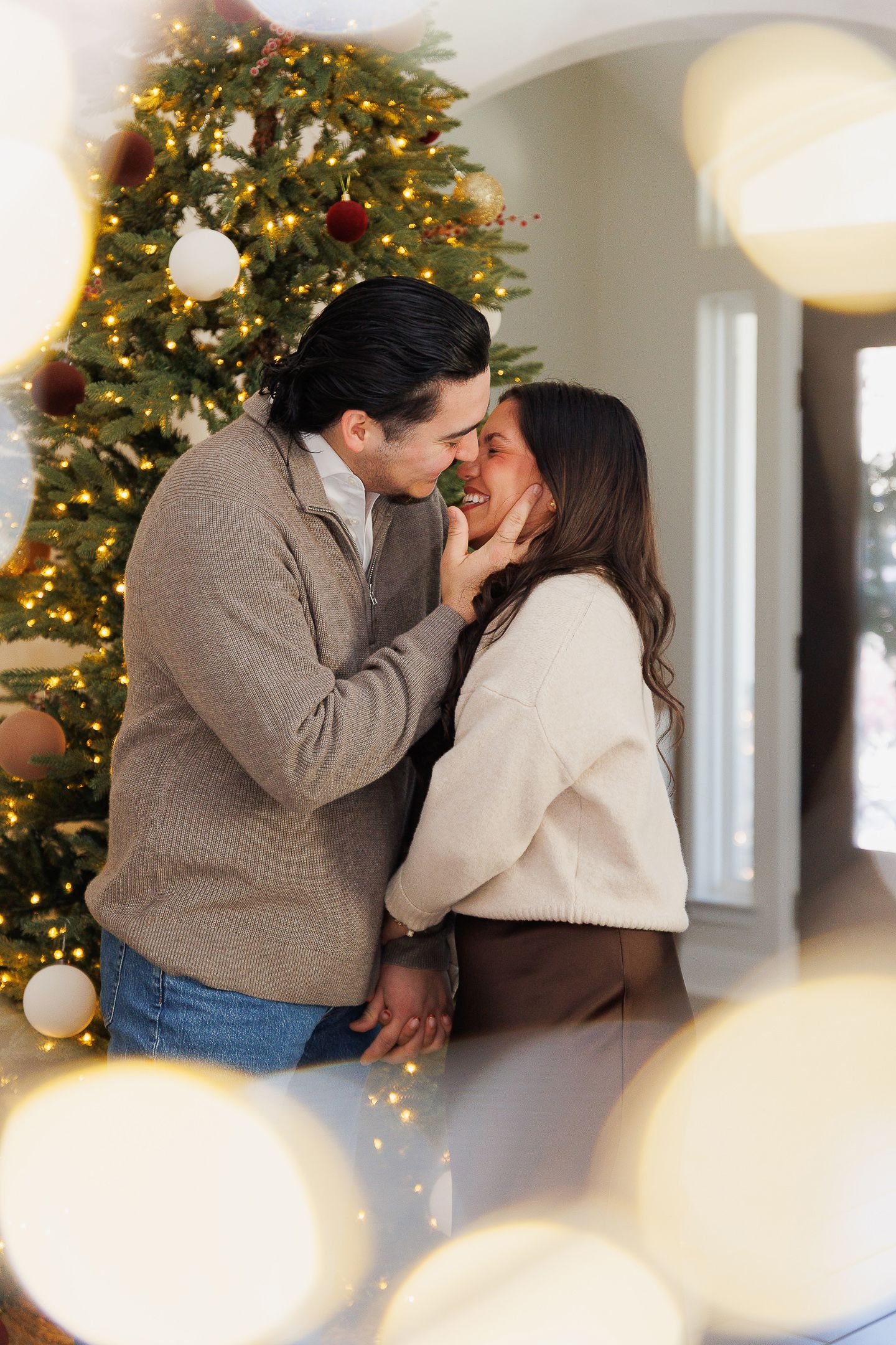 A couple shares a romantic moment in front of a decorated Christmas tree in a bright interior setting.