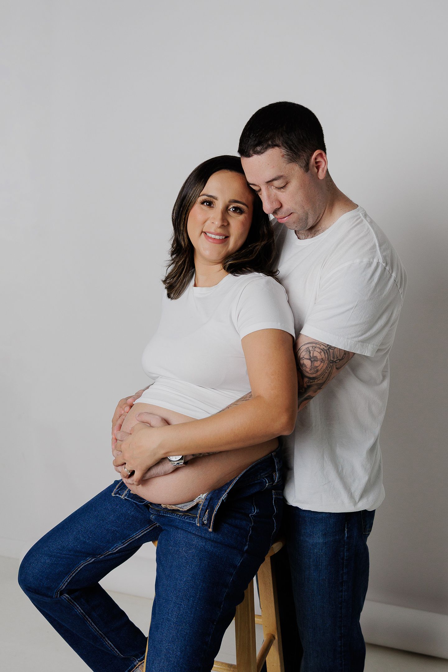 A couple in white shirts and jeans share tender moments during a maternity photoshoot against a neutral background.