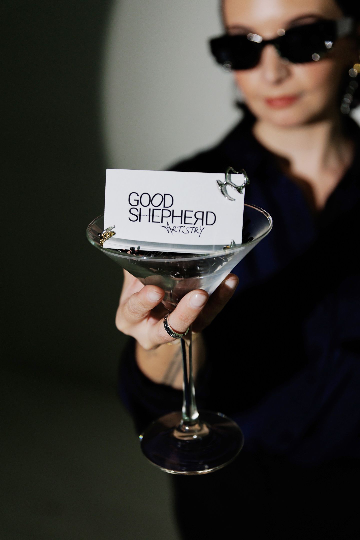 A stylish martini glass held up displaying a 'Good Shepherd' business card against a dark background.