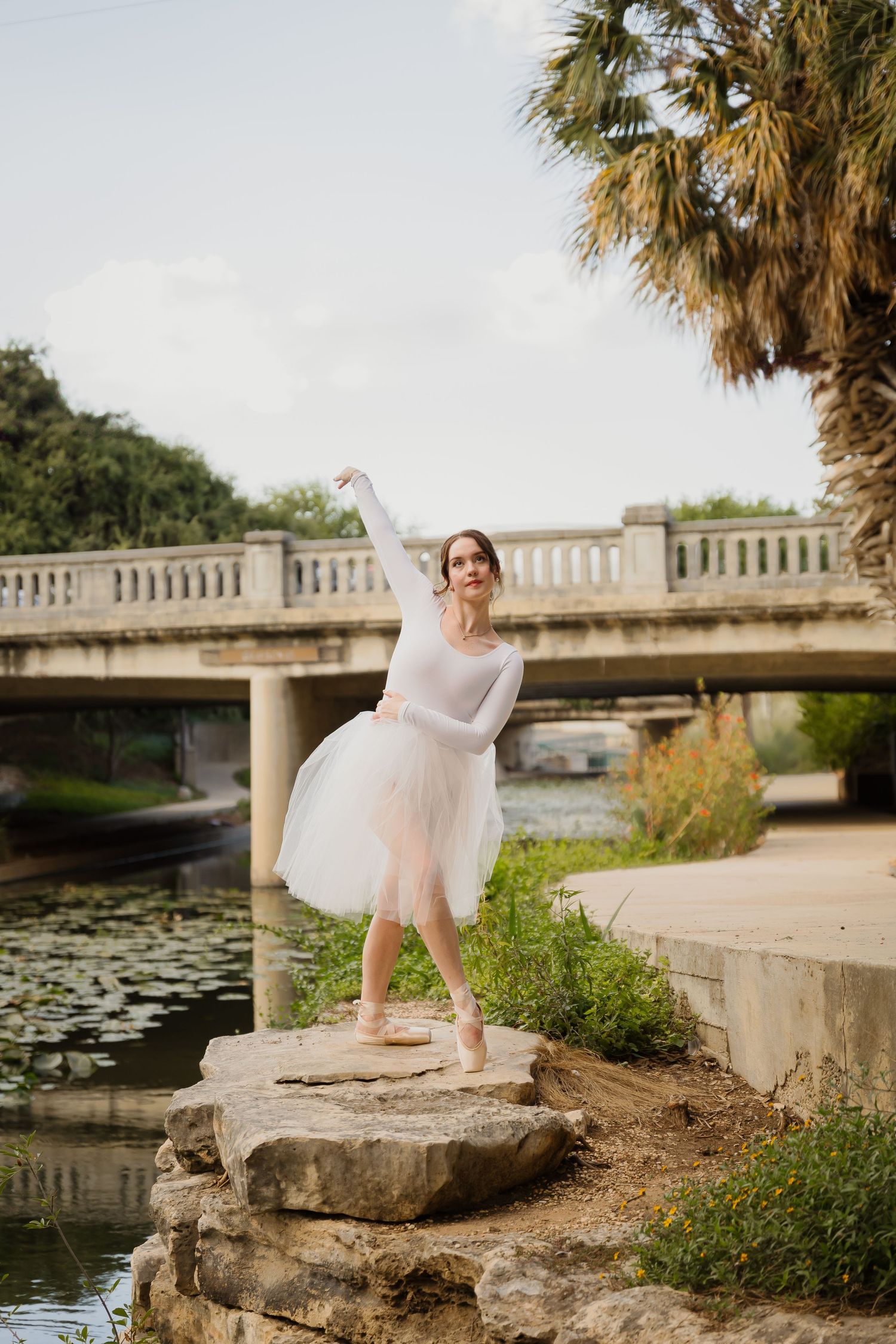 A ballet dancer in a tulle dress strikes a graceful pose on a rock beside a creek with a bridge in the background.