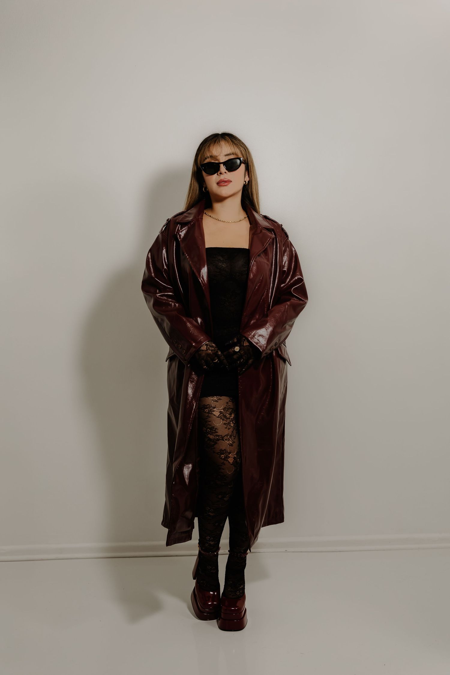 Fashion model wearing burgundy patent leather coat, black dress and platform boots against white background.