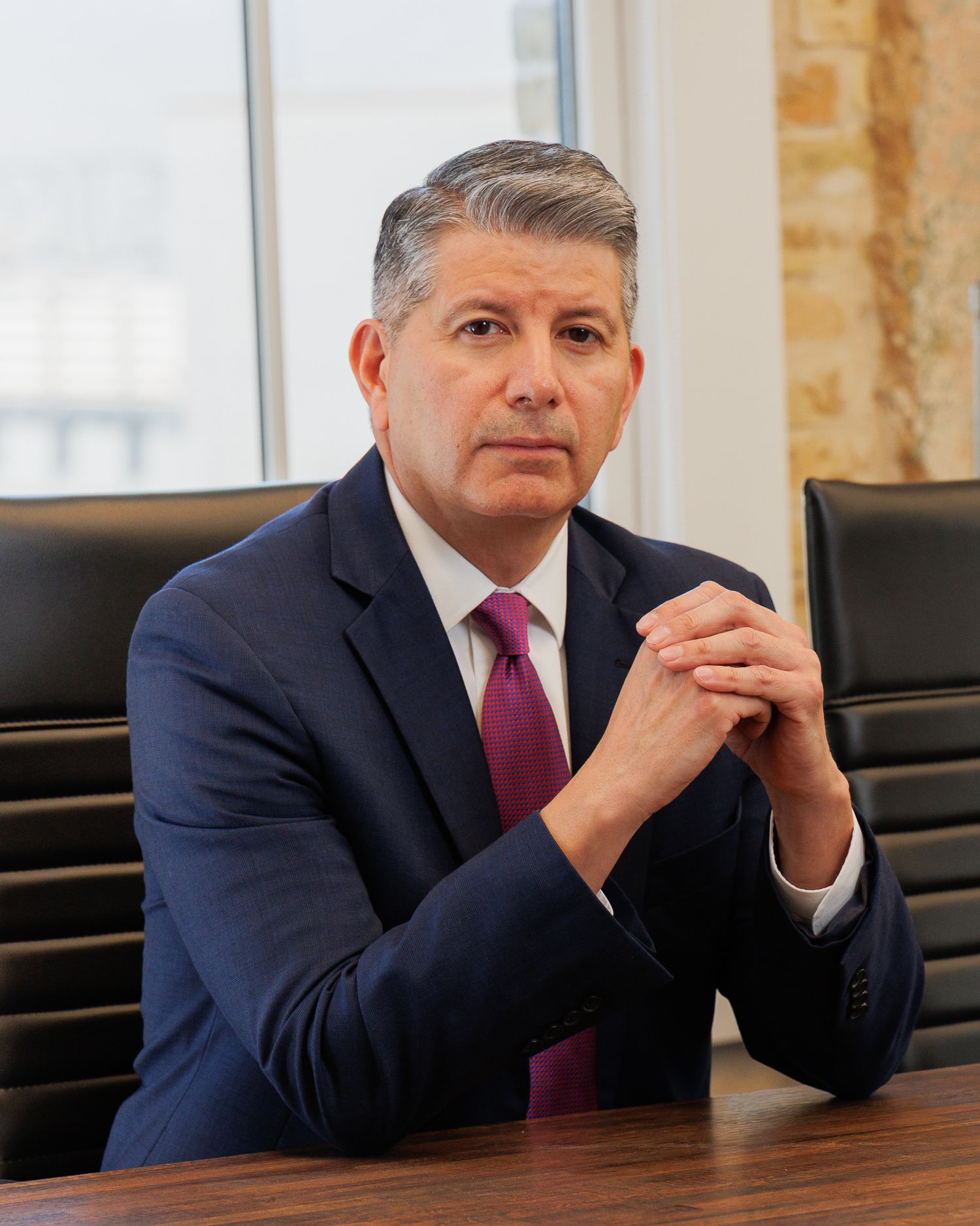 Professional businessman in navy suit and purple tie seated at conference table with hands clasped.
