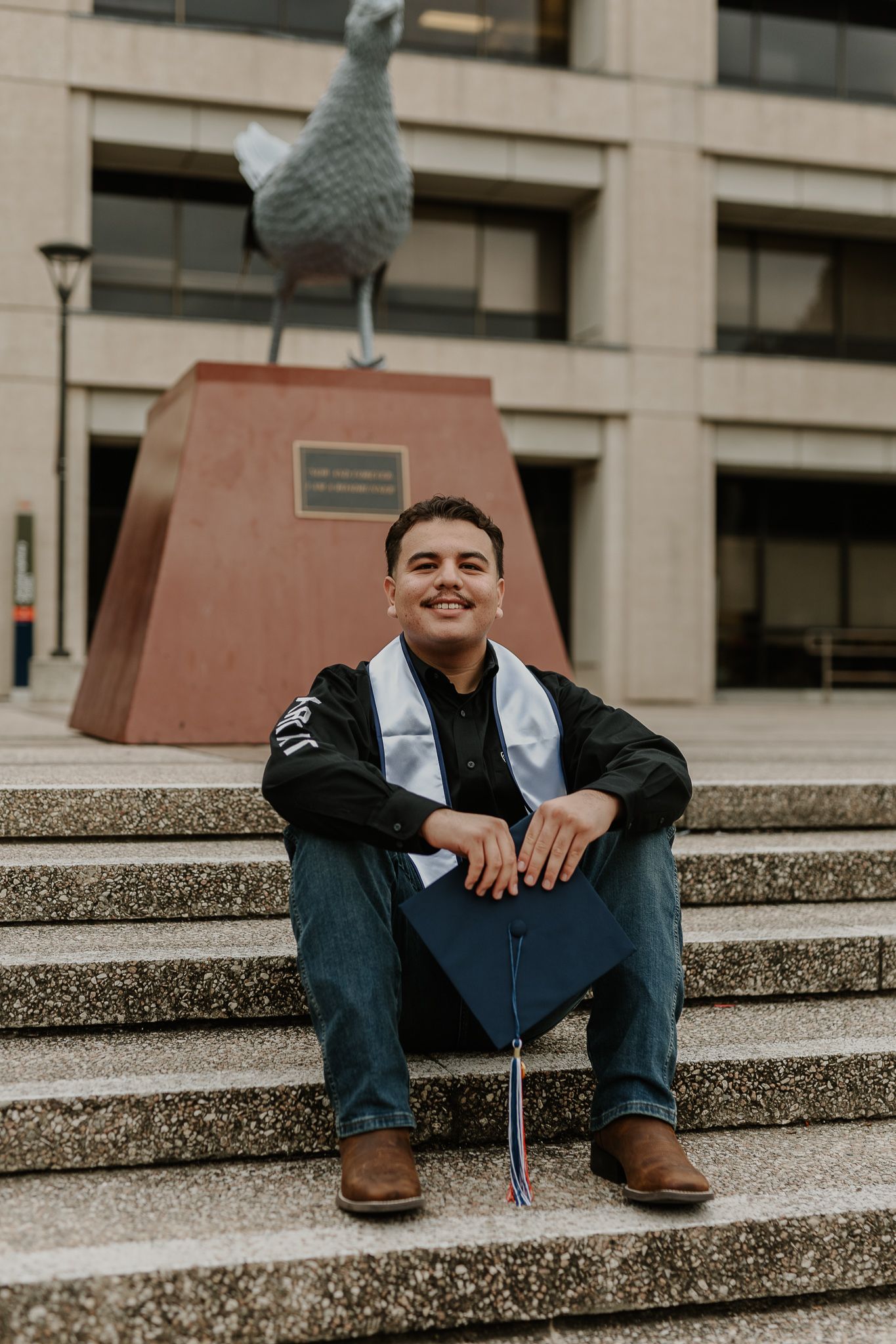 A graduate sitting on concrete steps with a large bird sculpture behind them.