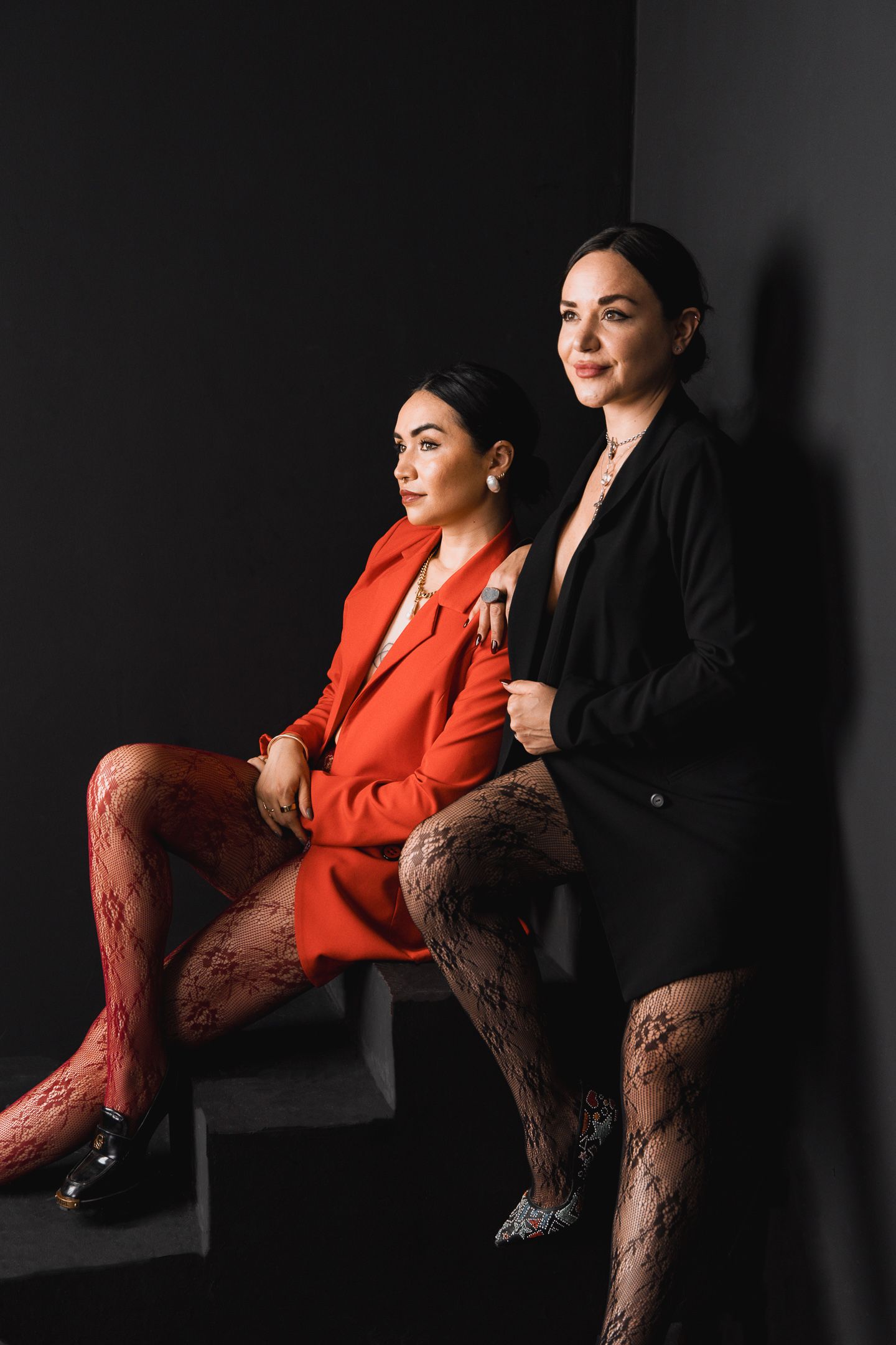 Two stylish women in lace tights pose on dark stairs, one in red blazer, one in black blazer.