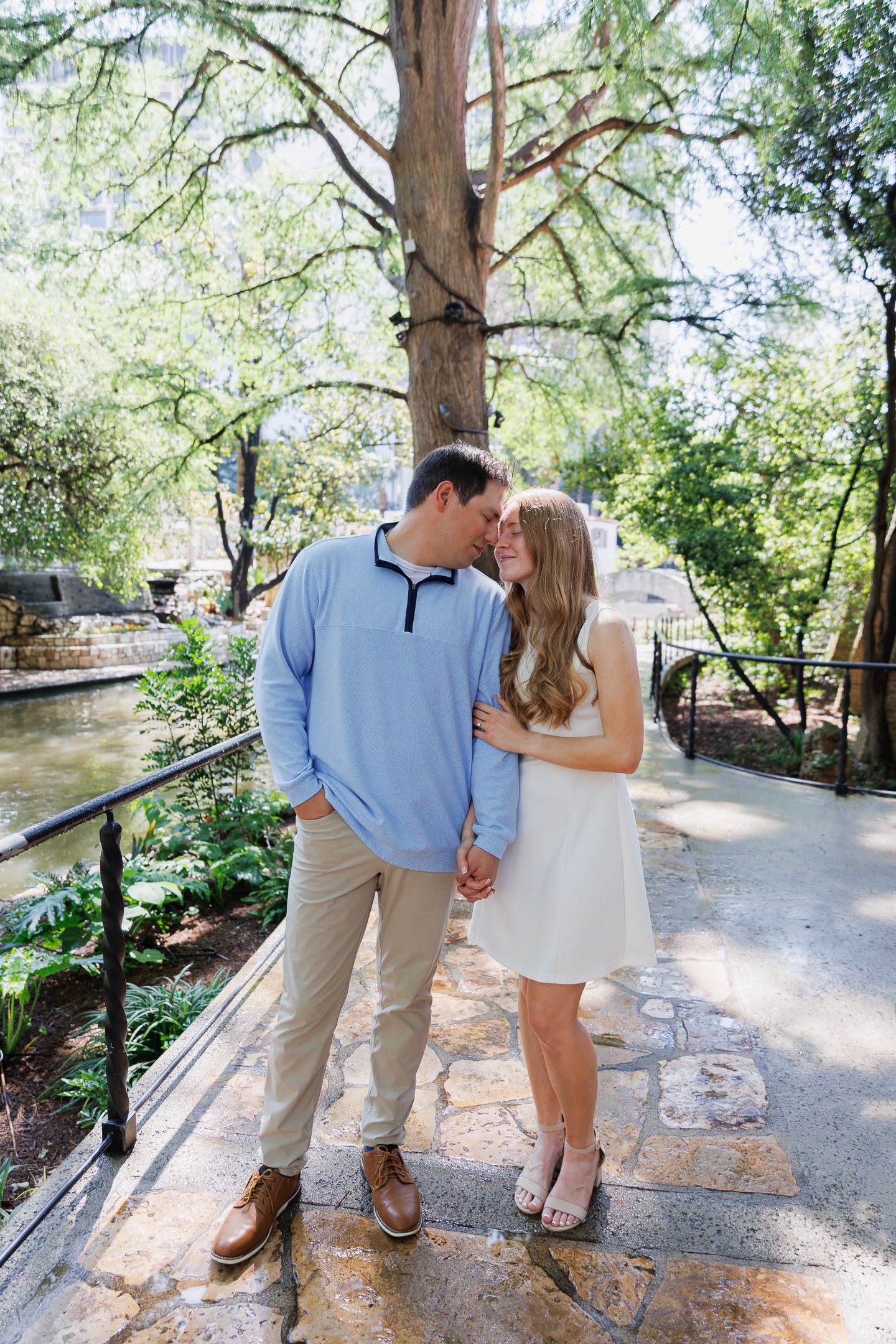A couple shares a romantic moment on a stone path surrounded by lush greenery near a river.