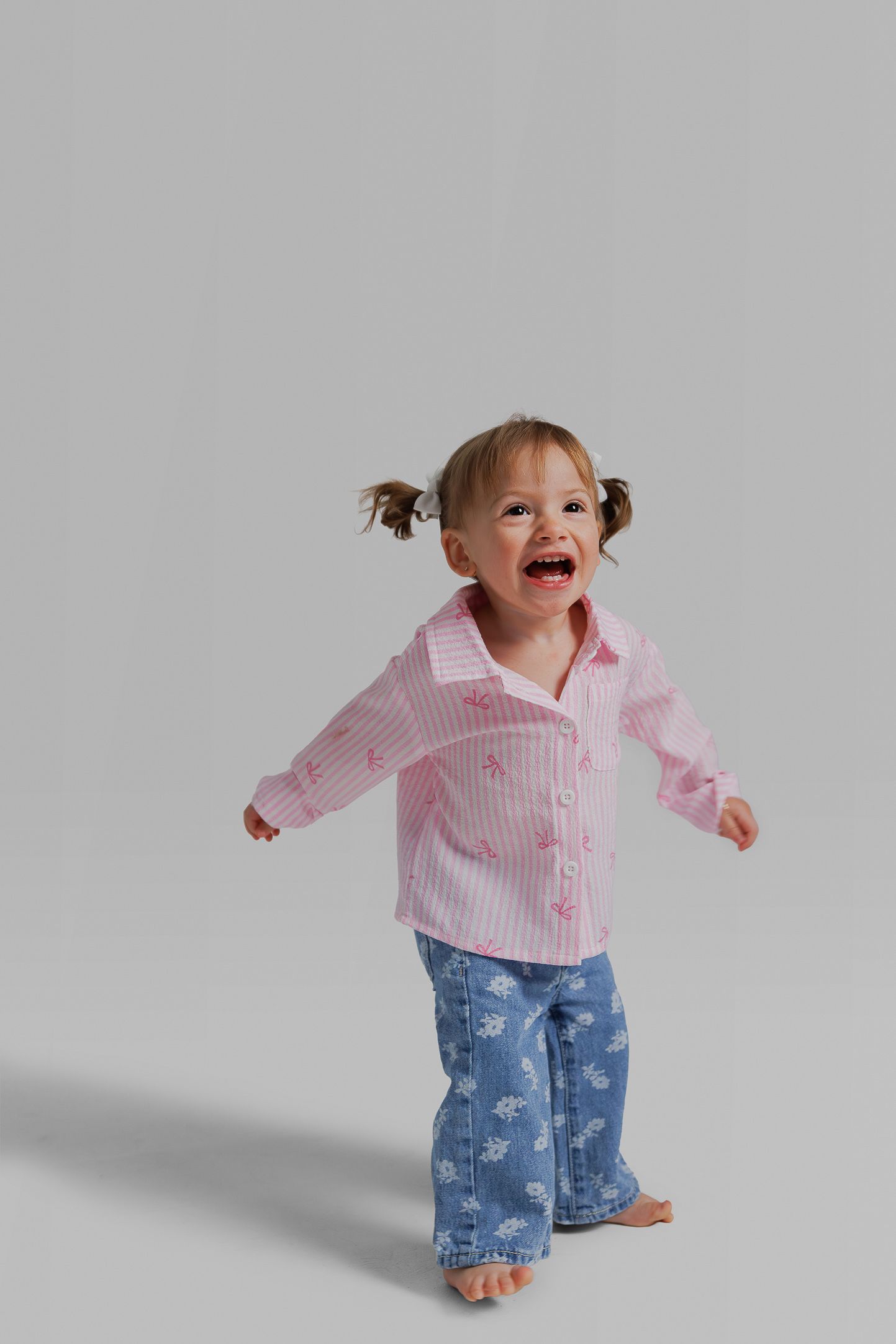 A toddler in a pink striped shirt and denim pants jumps and plays with excitement on a light gray background.