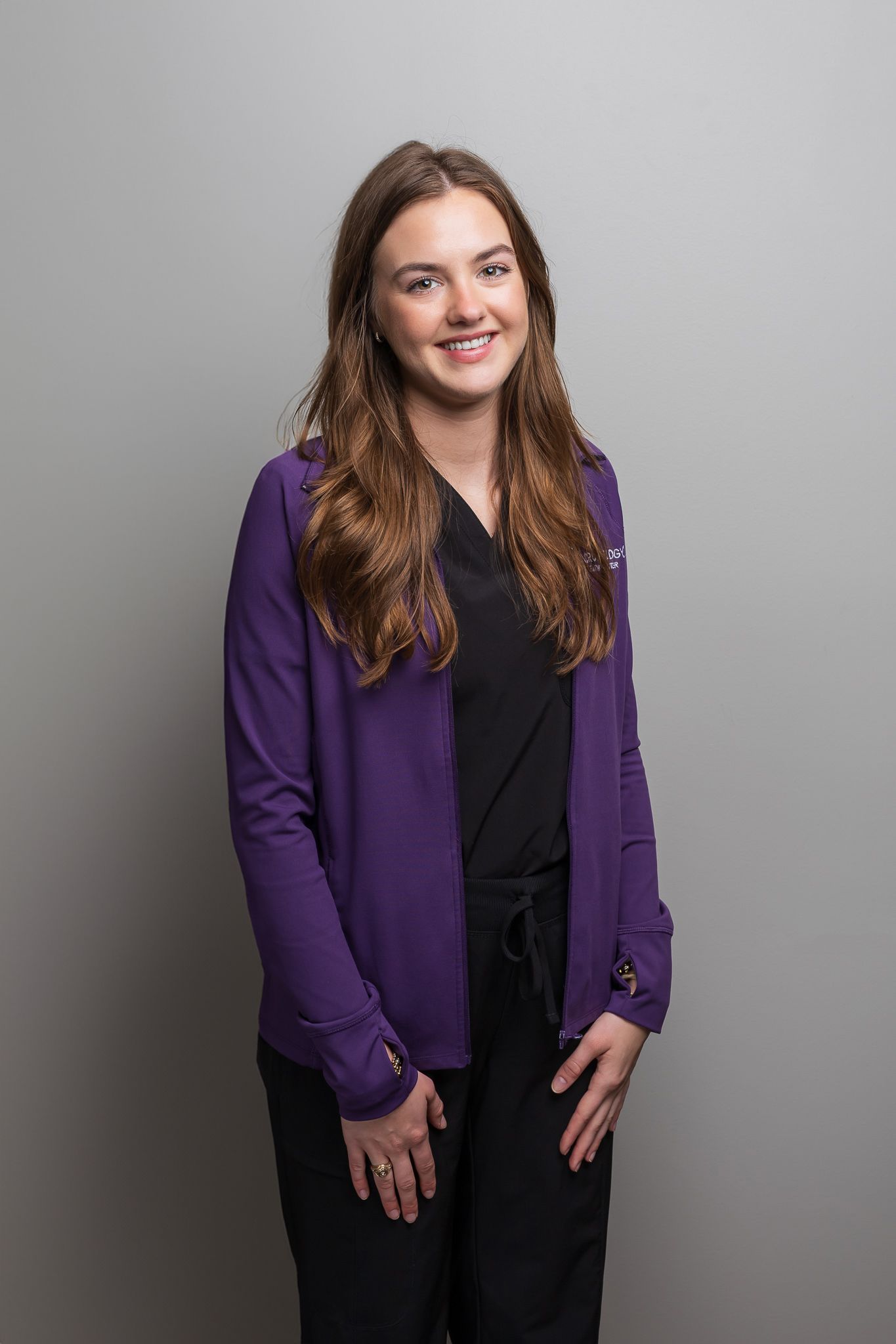 Healthcare worker wearing purple cardigan and black scrubs poses against grey background.