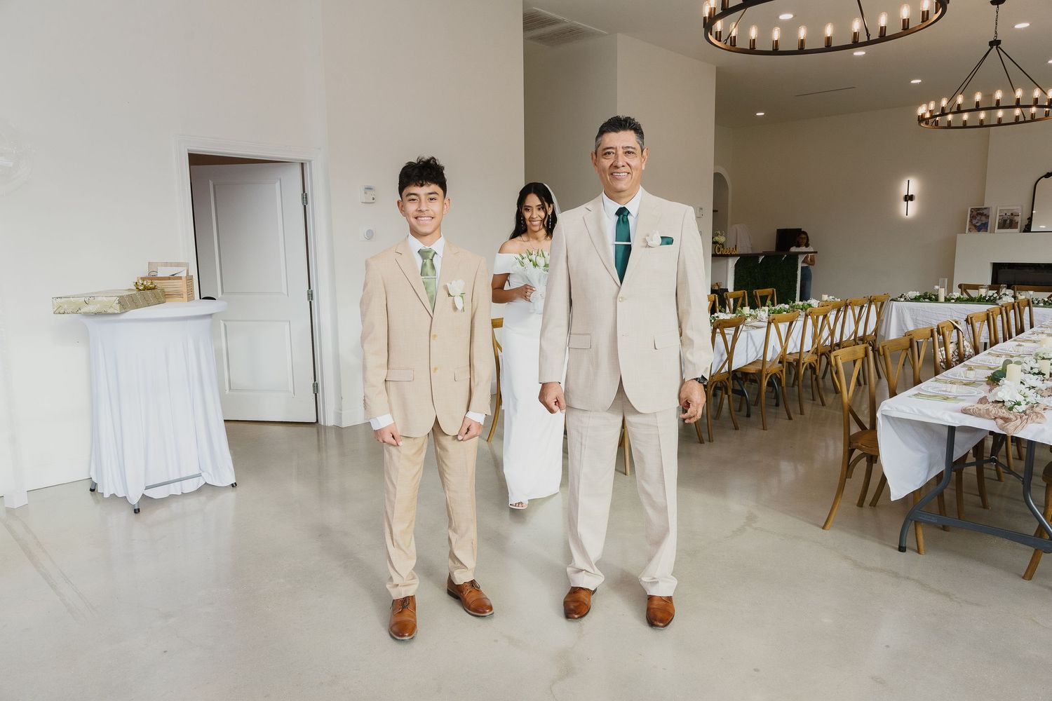 Three people in formal attire standing in a modern white reception venue with elegant table settings.