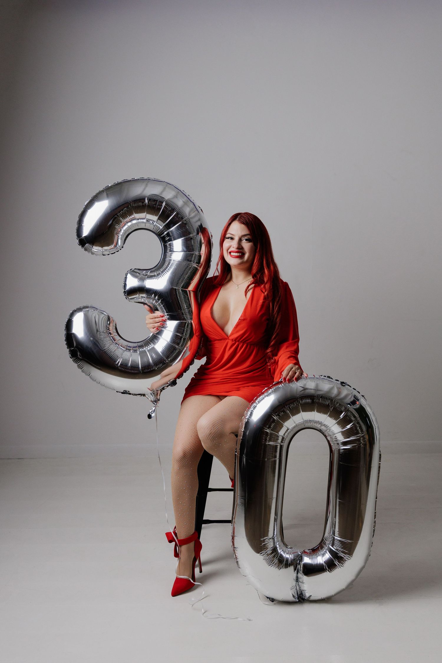 A person in a red dress poses with large silver number balloons against a gray backdrop for a 30th birthday photoshoot.