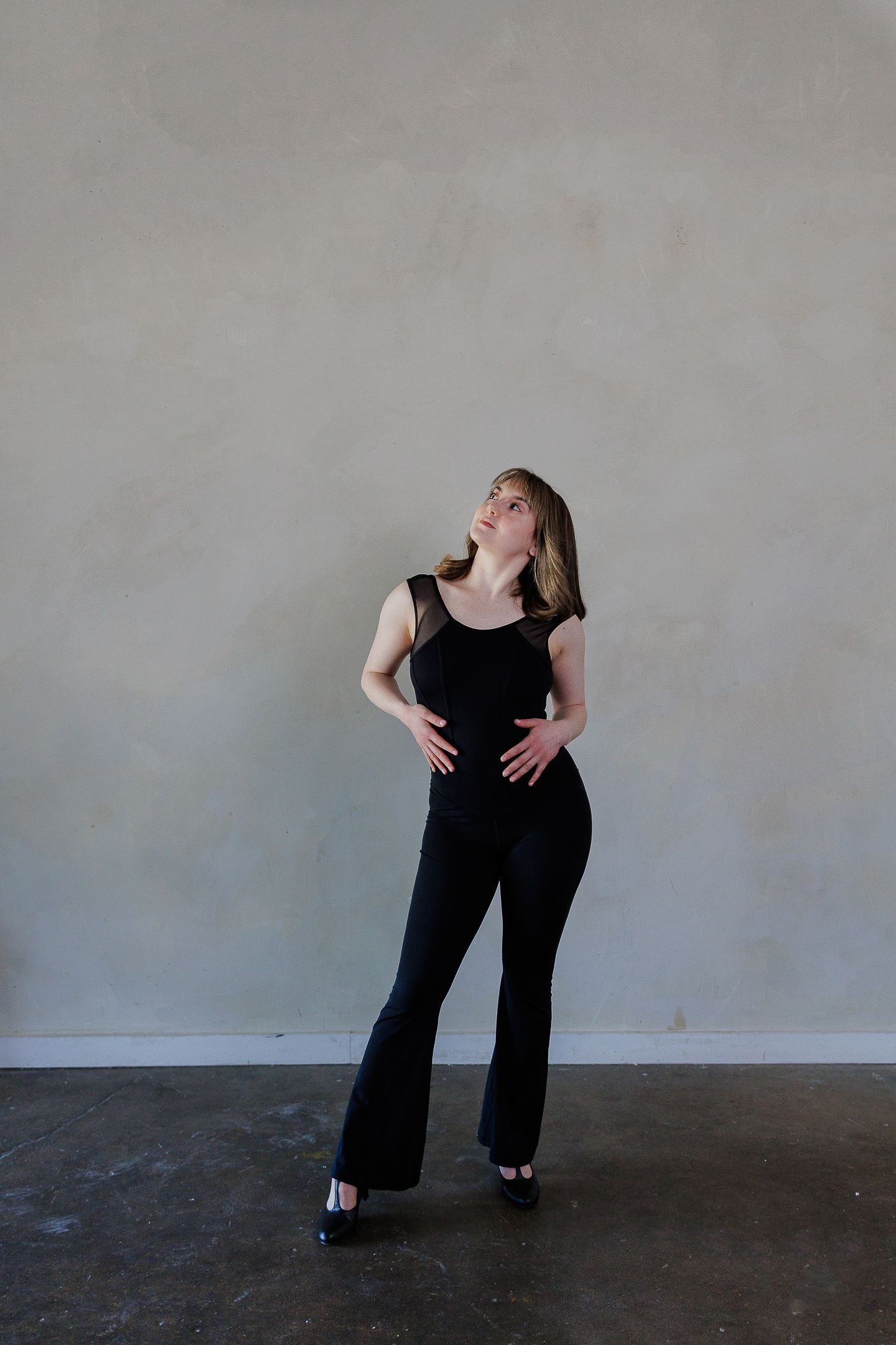 A figure in black clothing strikes dynamic dance poses against a plain gray wall.