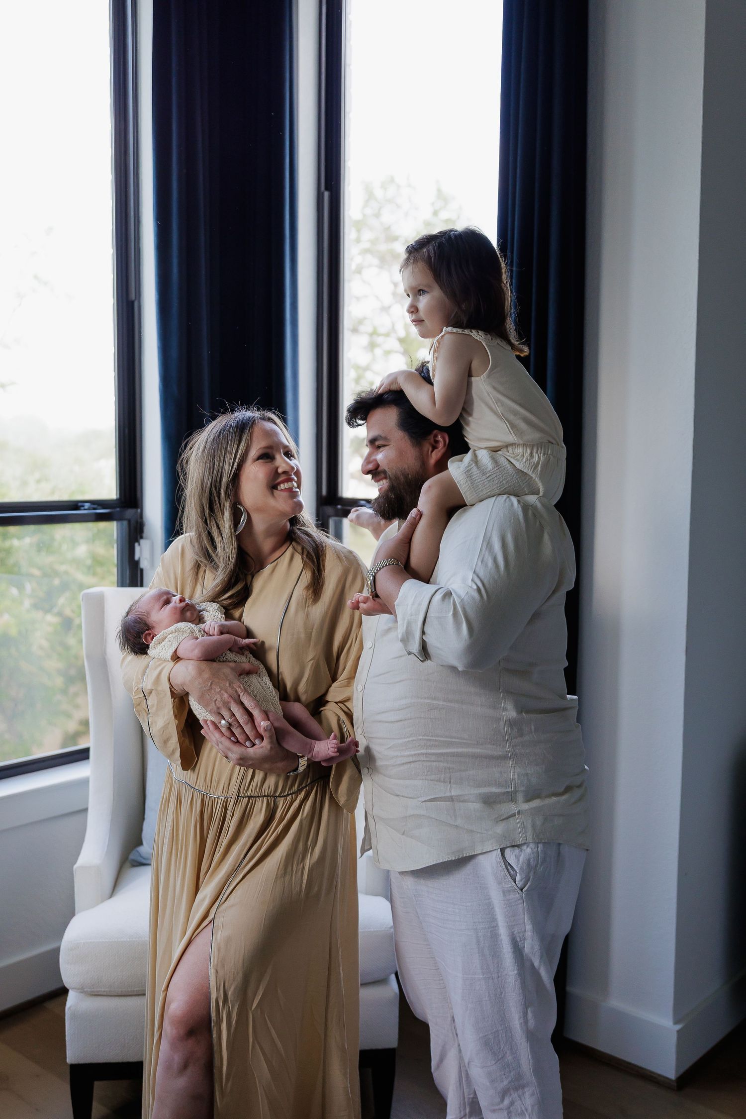 A couple in white and beige clothing embraces lovingly by large windows in a bright modern room.