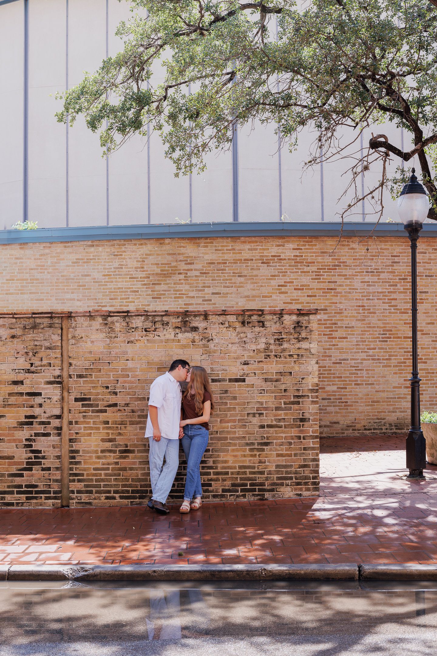 Couple sharing a romantic kiss against a rustic brick wall on a sunny day in an urban setting.