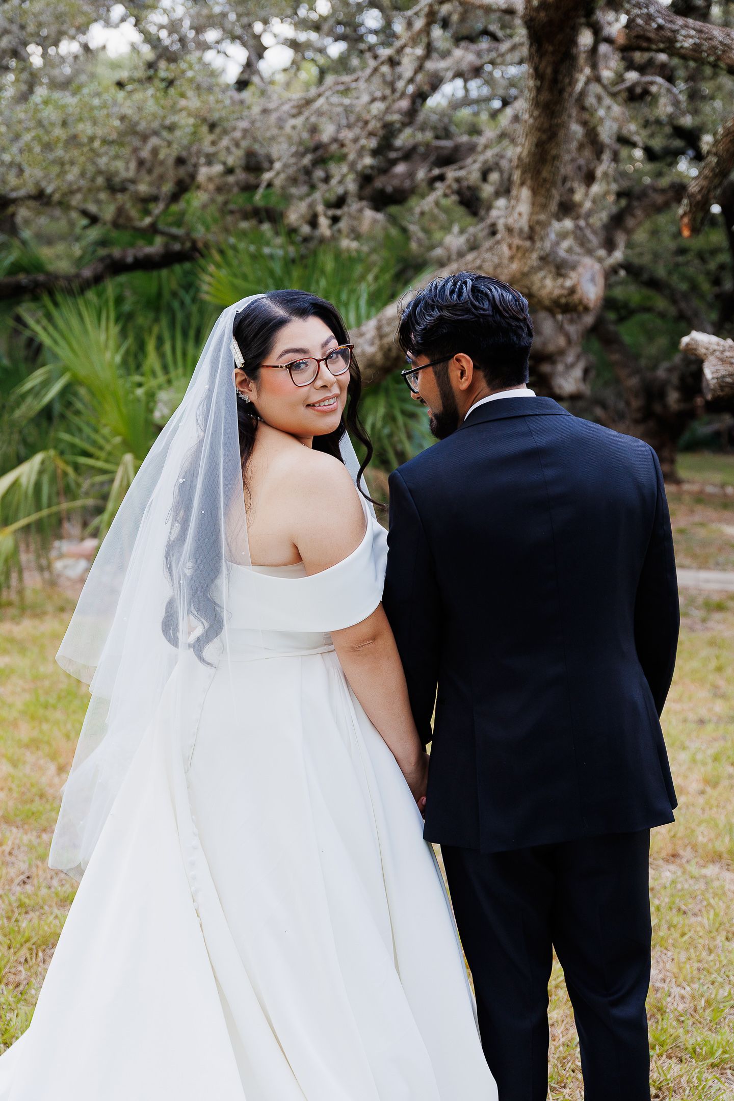 A couple in wedding attire stands together outdoors with blooming trees in the background.