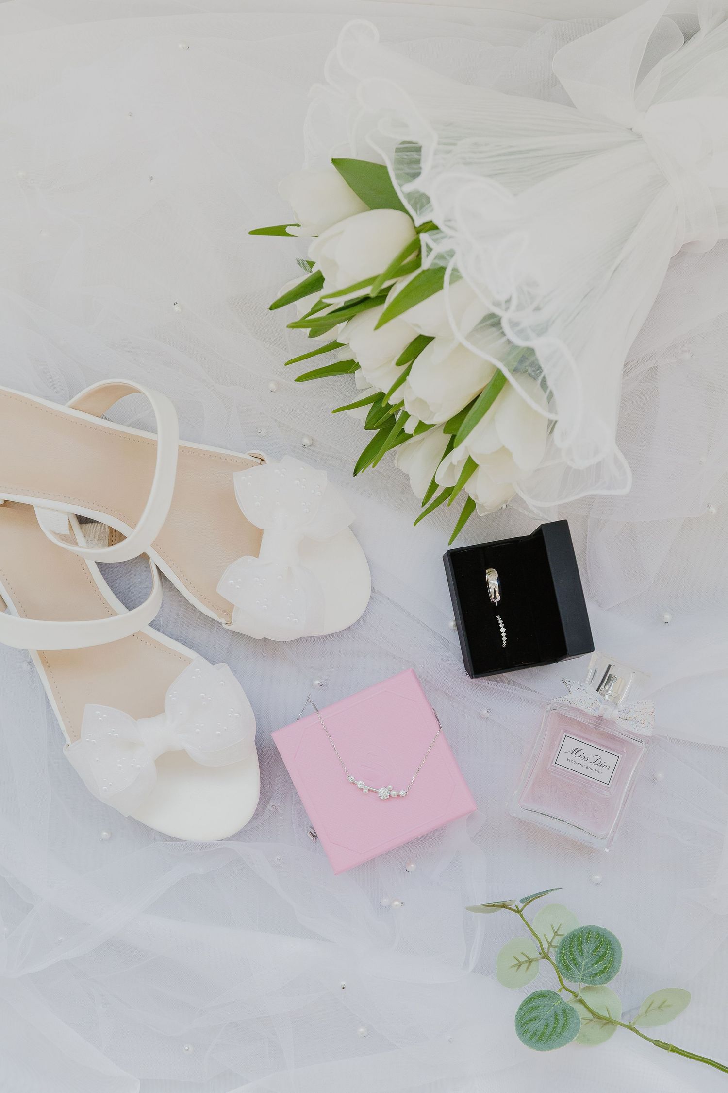 Wedding details flatlay with white tulips, heels, rings, and perfume on tulle fabric.