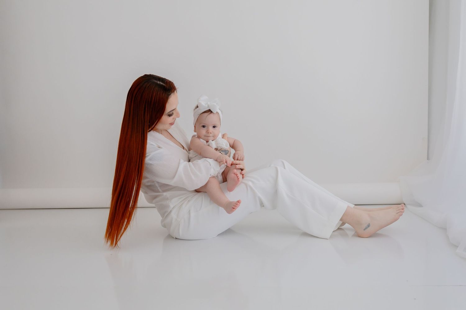 Parent with long red hair in white outfit sitting on floor playing with baby.