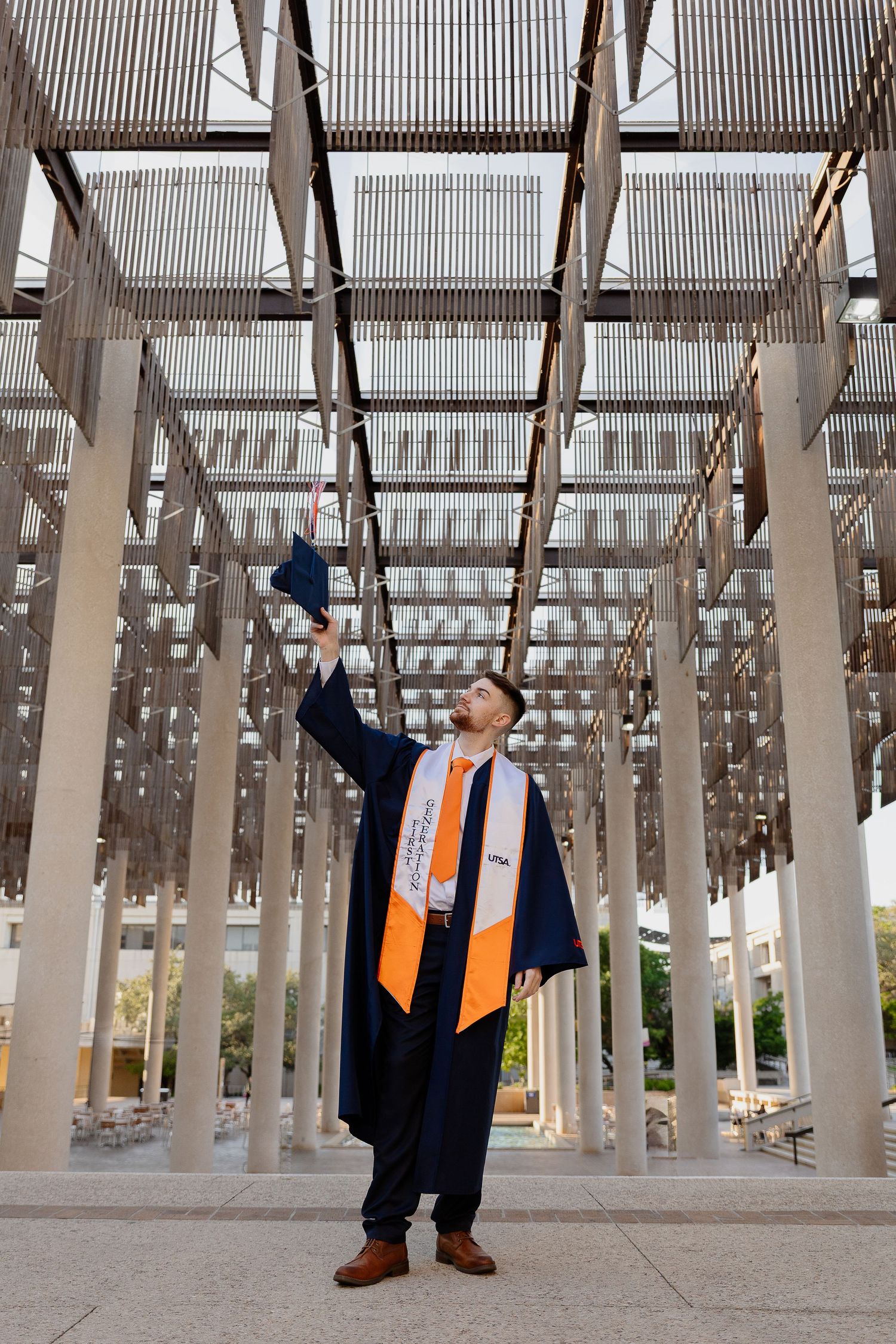 Graduate in cap and gown throws graduation cap under architectural pergola structure.