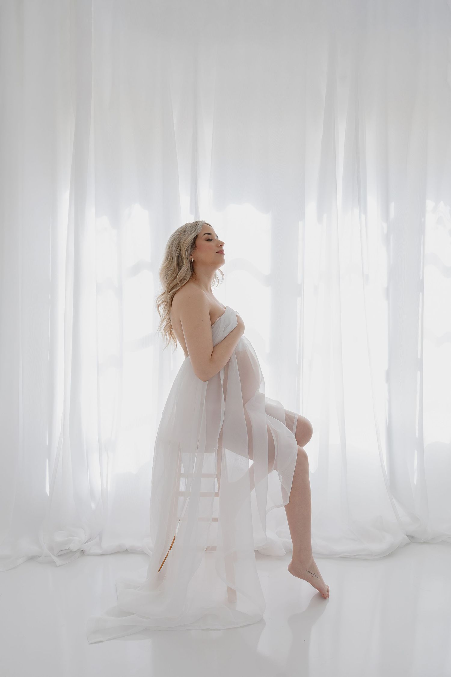 Artistic maternity portrait with flowing white fabric against bright window.