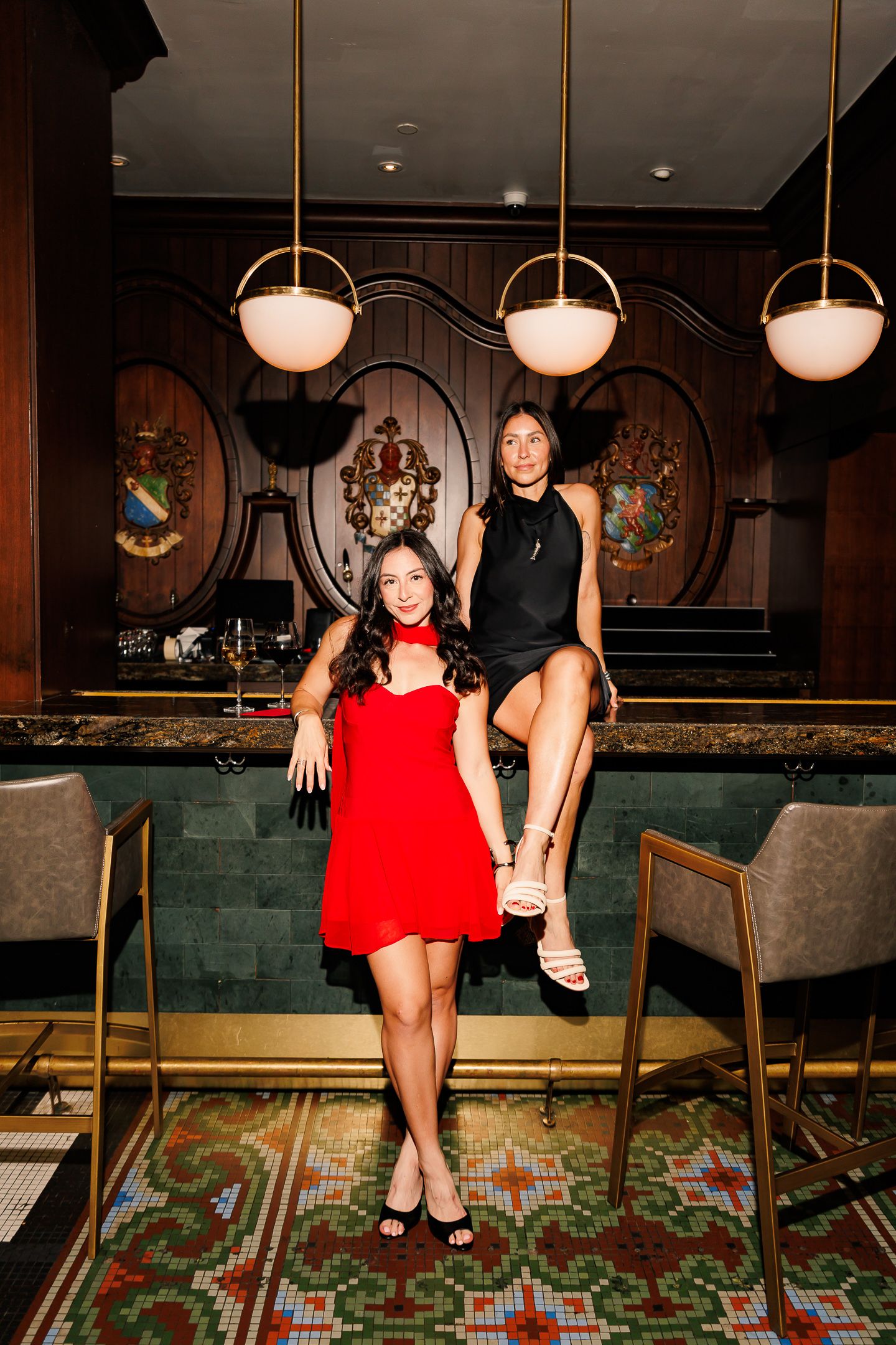 Two women in elegant dresses pose at an upscale bar, one in red standing, one in black seated on counter.