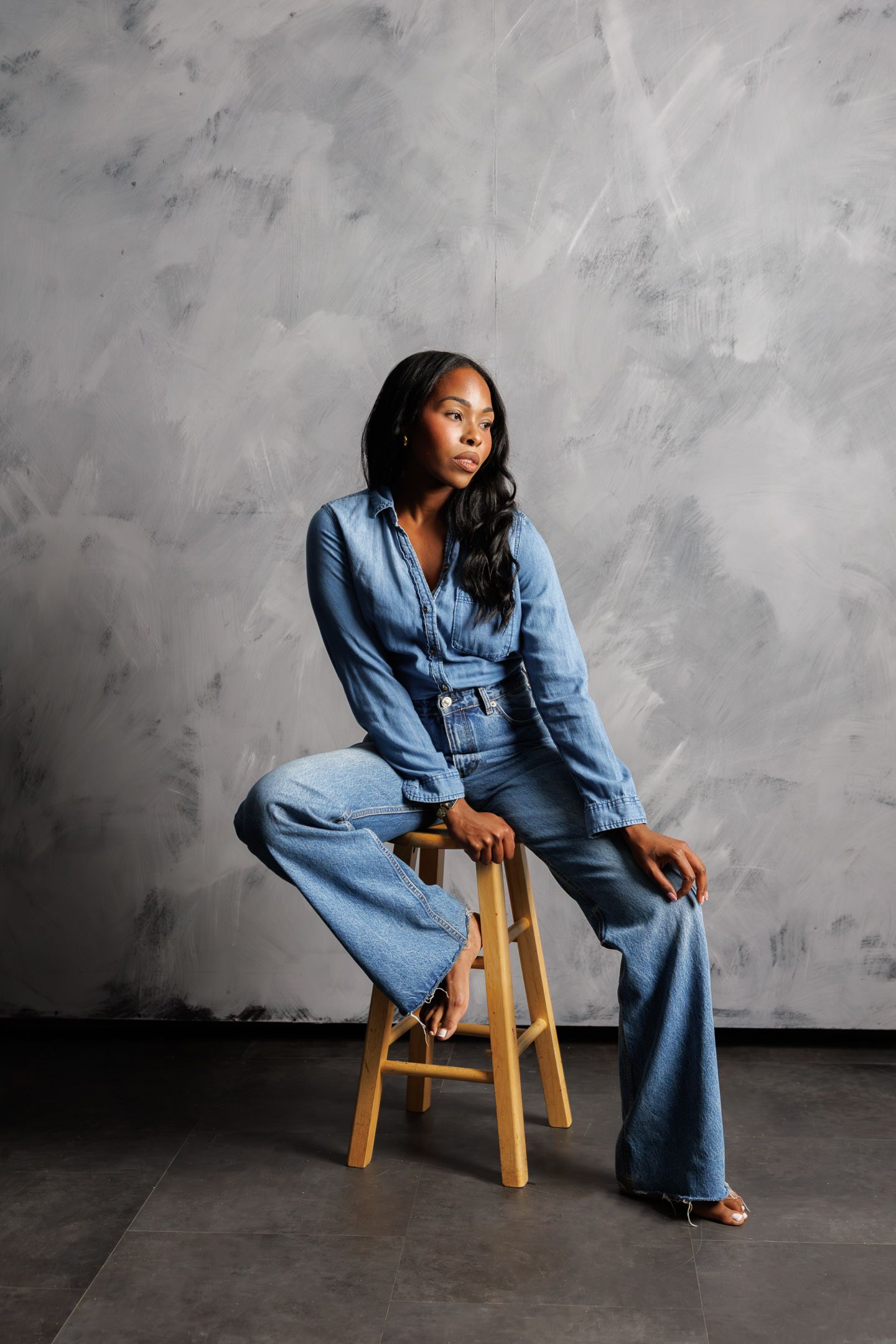 A woman in a denim jumpsuit sits on a wooden stool against a gray textured wall.