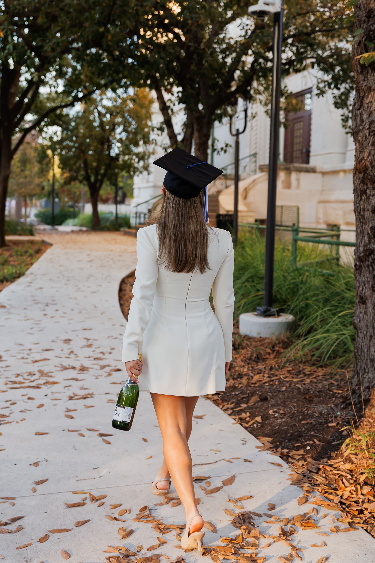 A figure in a white dress and black hat walks along a path covered in autumn leaves.