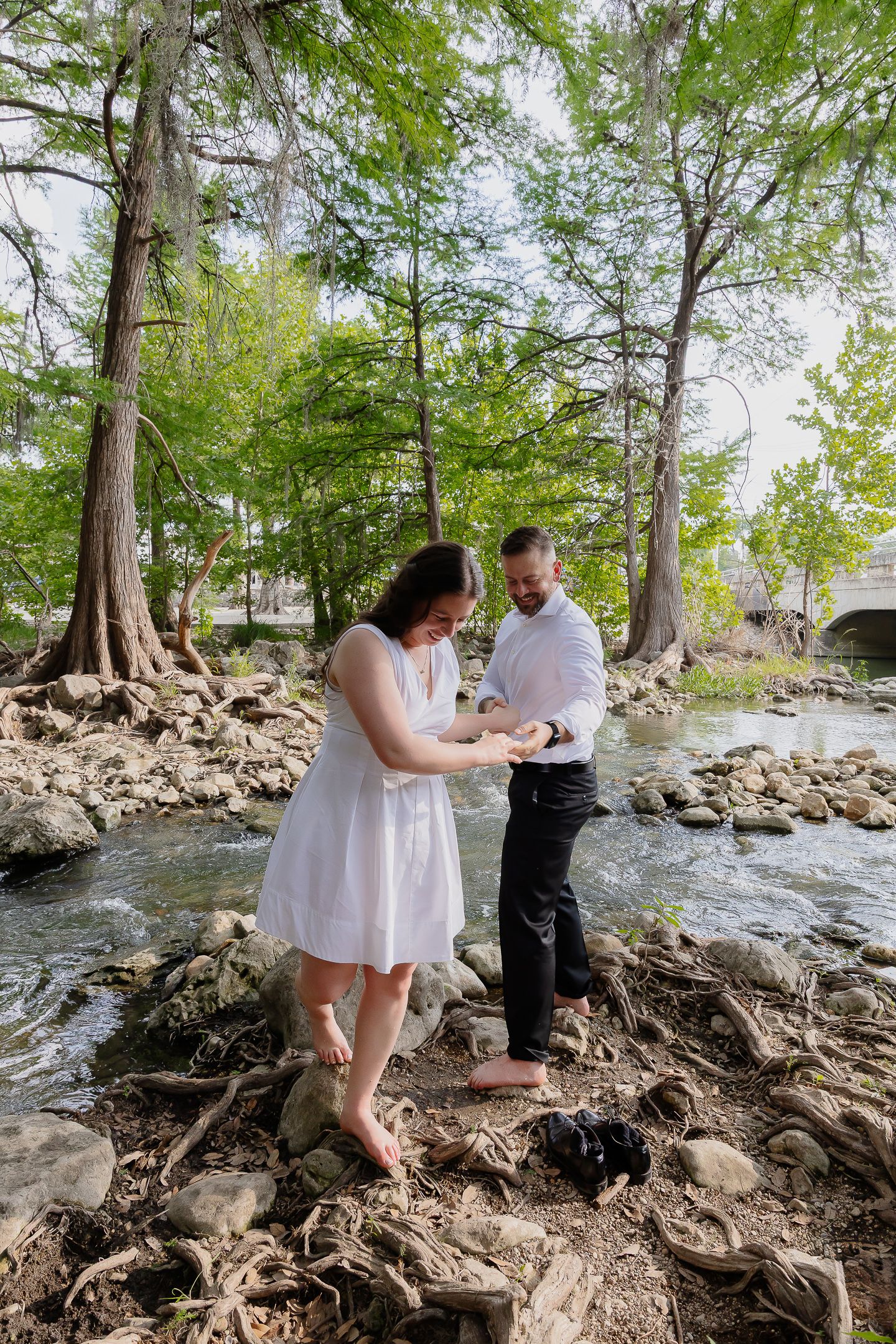 A couple shares a romantic moment among exposed tree roots and rocks along a scenic riverbank in a forest setting.
