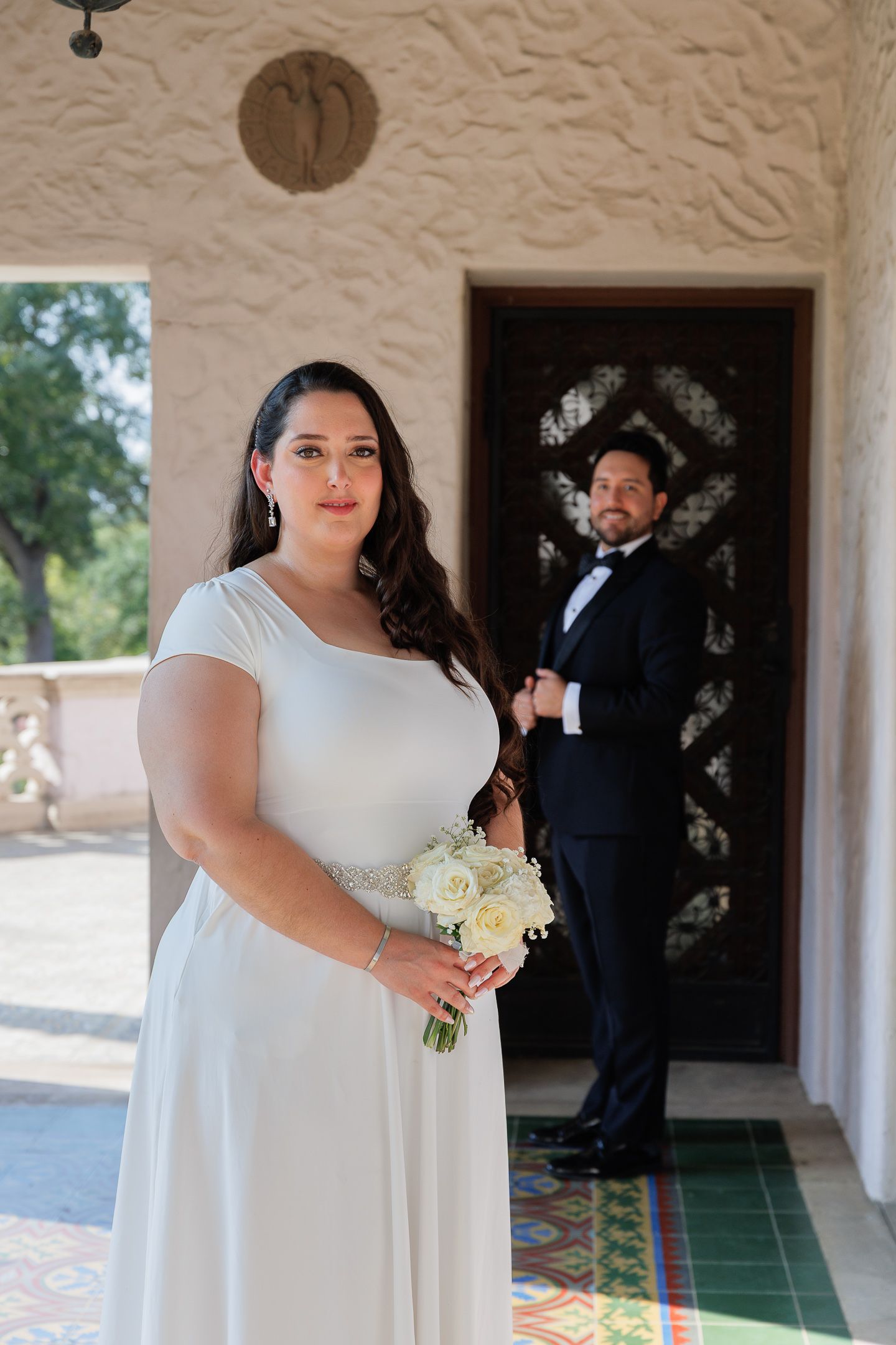 A bride in a classic white gown holds a white bouquet while standing in an elegant arched corridor.