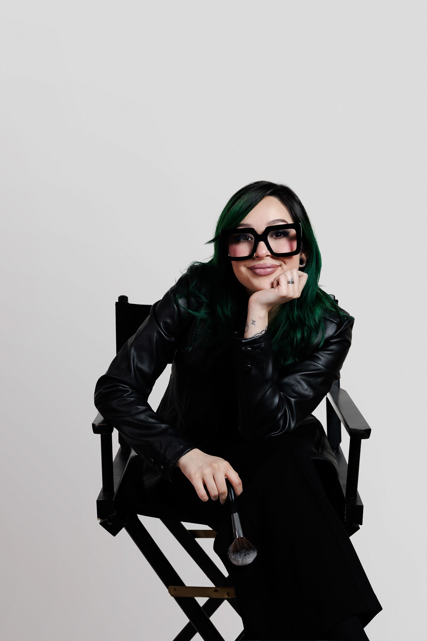 A person in black winter attire and dark glasses poses dramatically while seated in a director's chair against a gray backdrop.