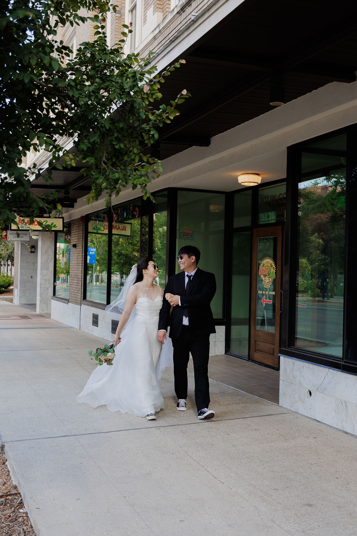 A couple in wedding attire poses together under a covered walkway with glass windows and concrete columns.
