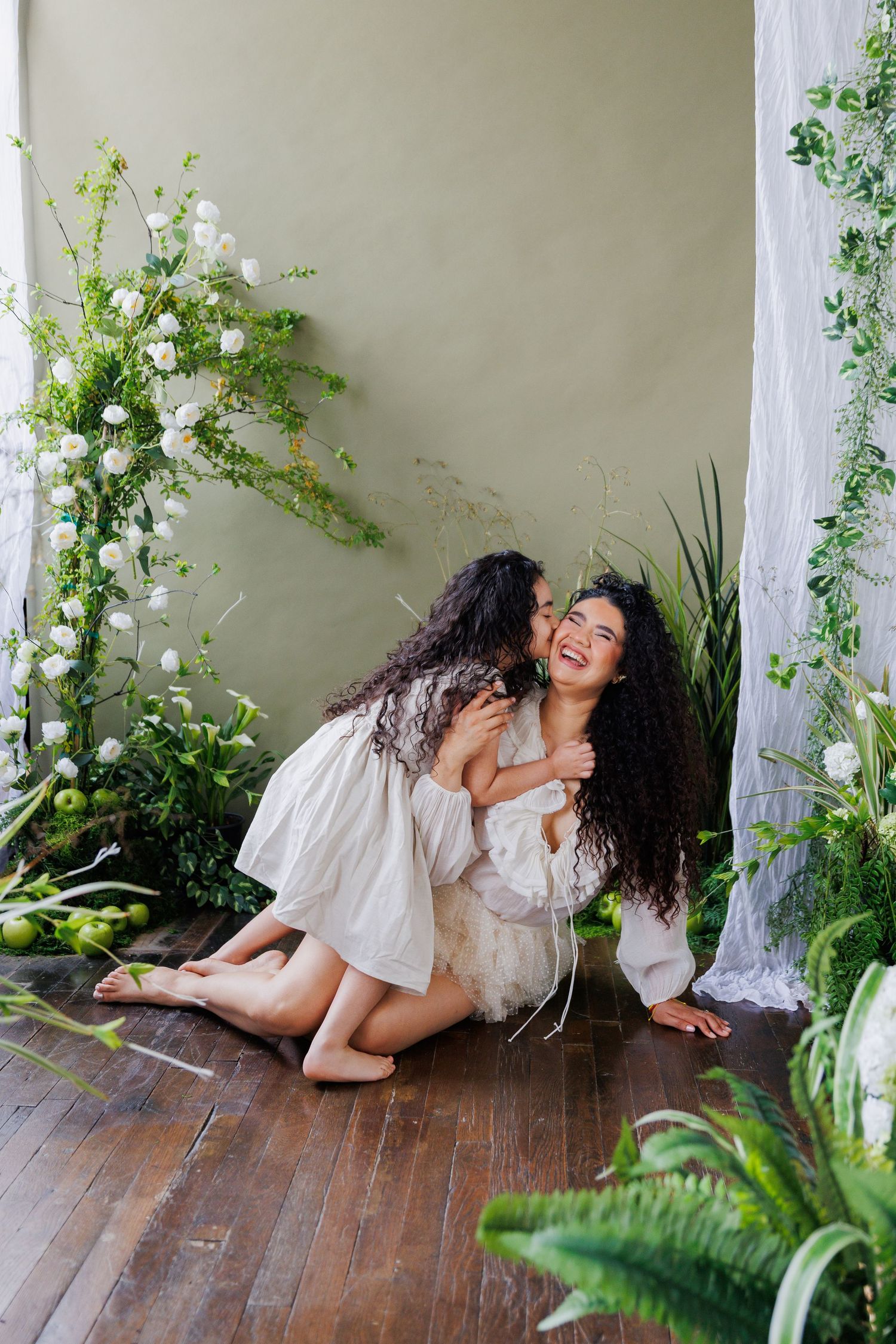 Two people in white outfits share intimate moments on a wooden floor surrounded by lush greenery and natural light.