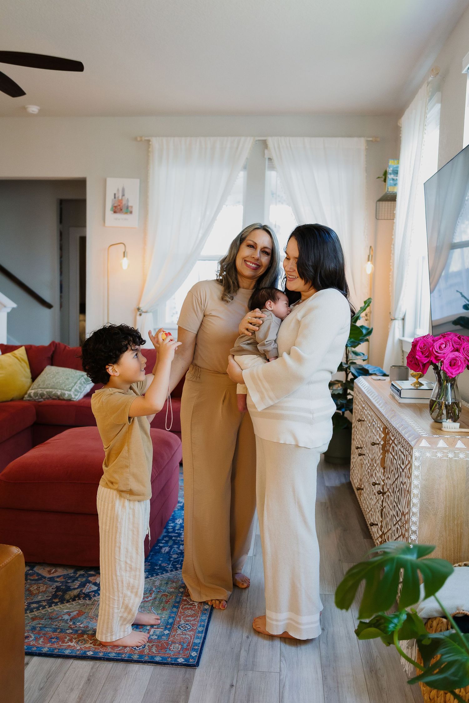 Three people having a casual conversation in a bright living room with red couch and decorative curtains.