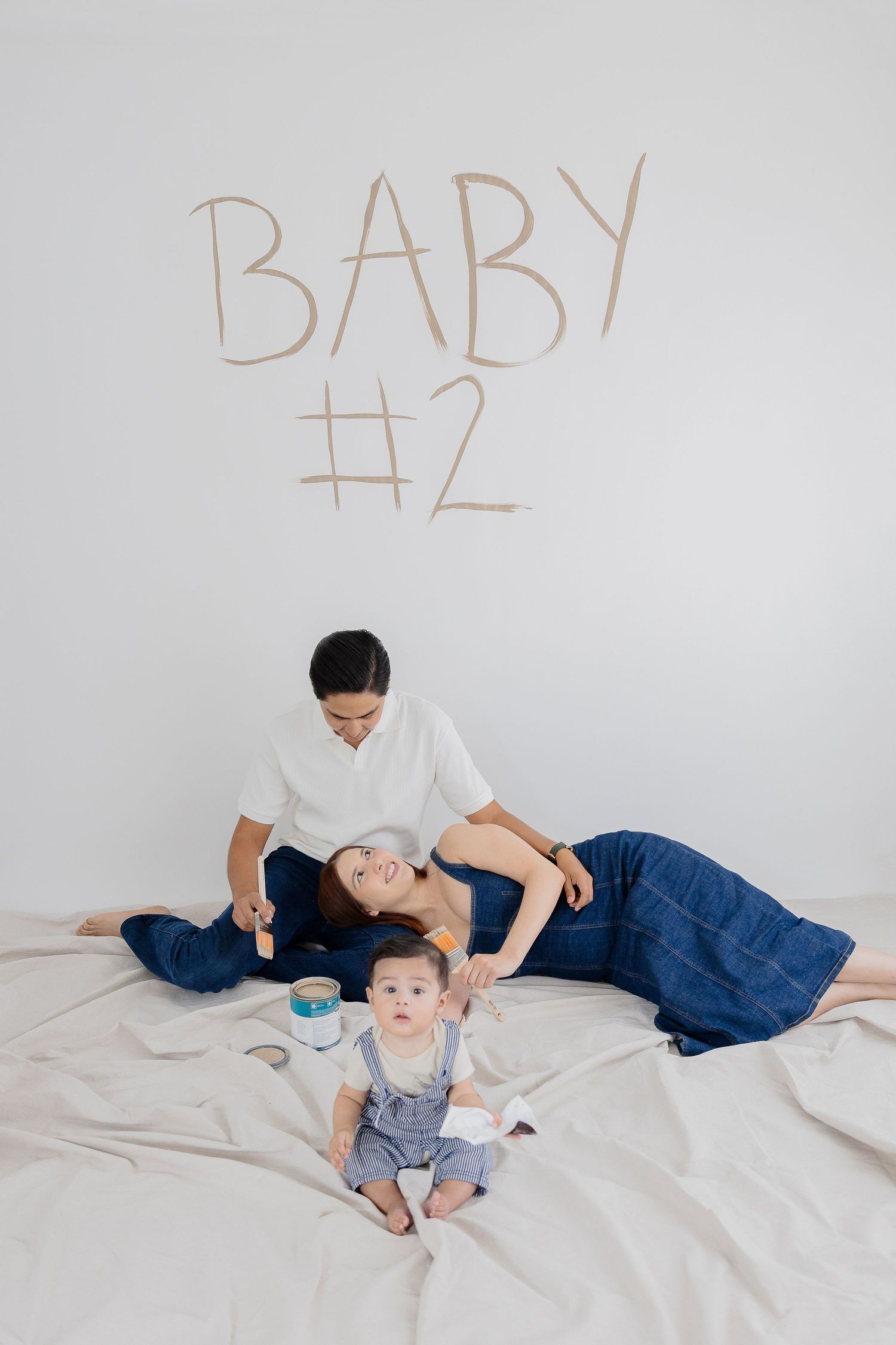 Family sits on white bedding against wall with 'BABY #2' text written above them for pregnancy announcement photo.