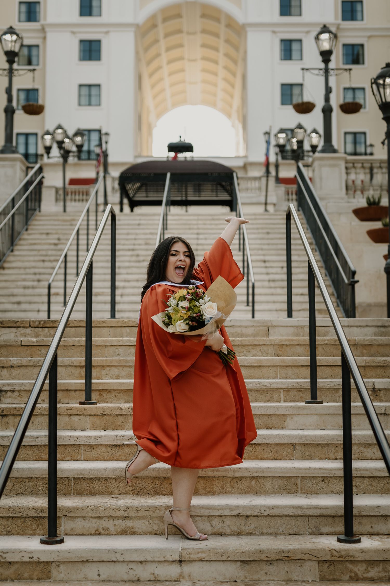 Graduate celebrating with bouquet and diploma on ornate stone staircase.