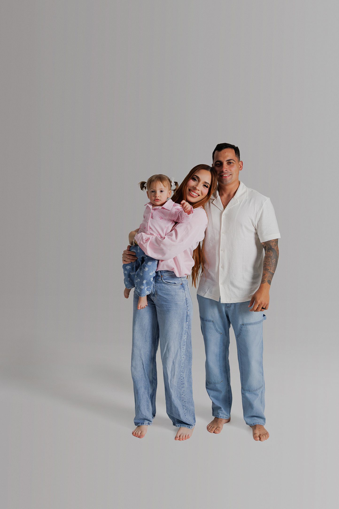 Family wearing casual denim and white outfits poses together against gray background in studio setting.