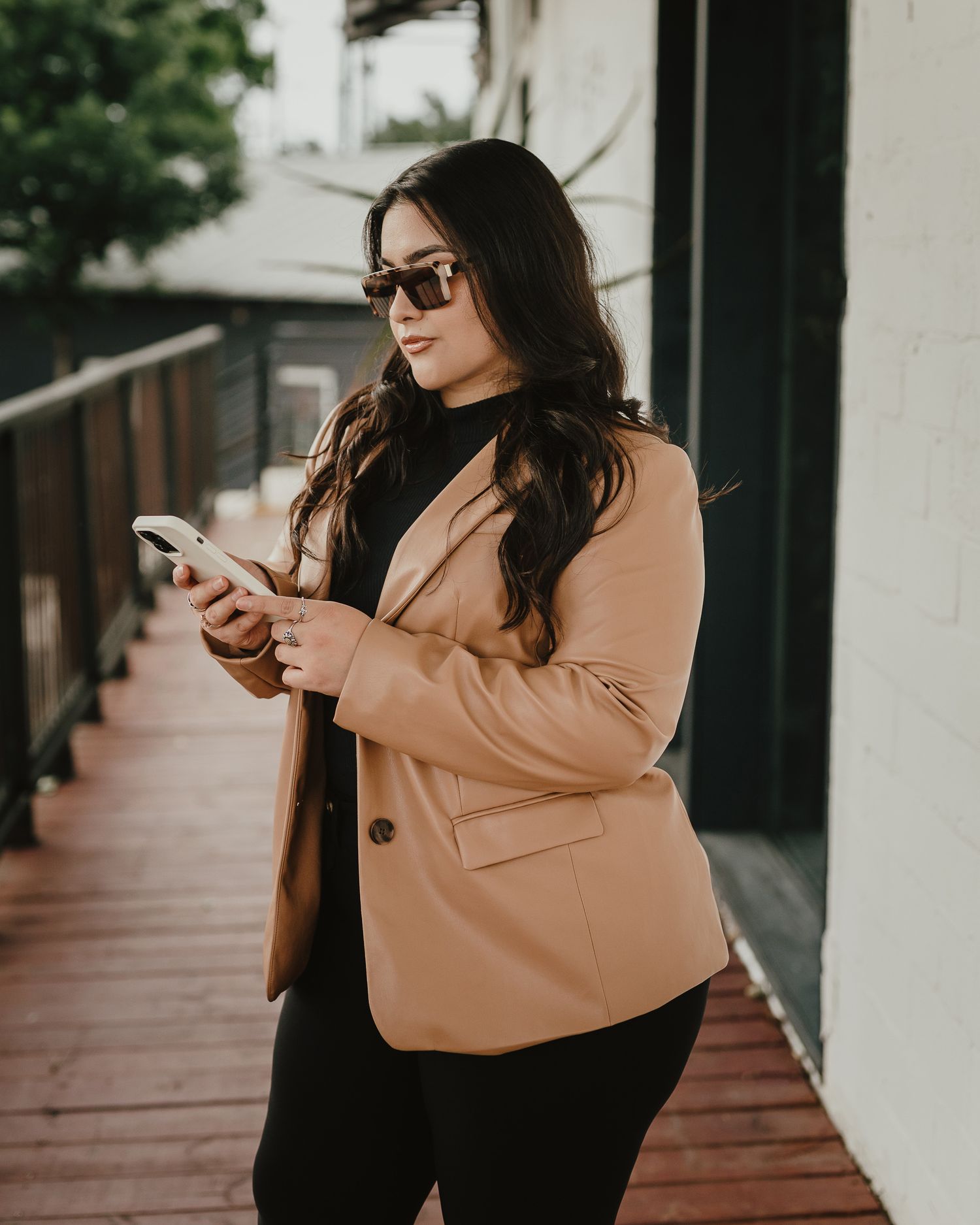 Business professional in tan blazer checks phone while standing on wooden deck.