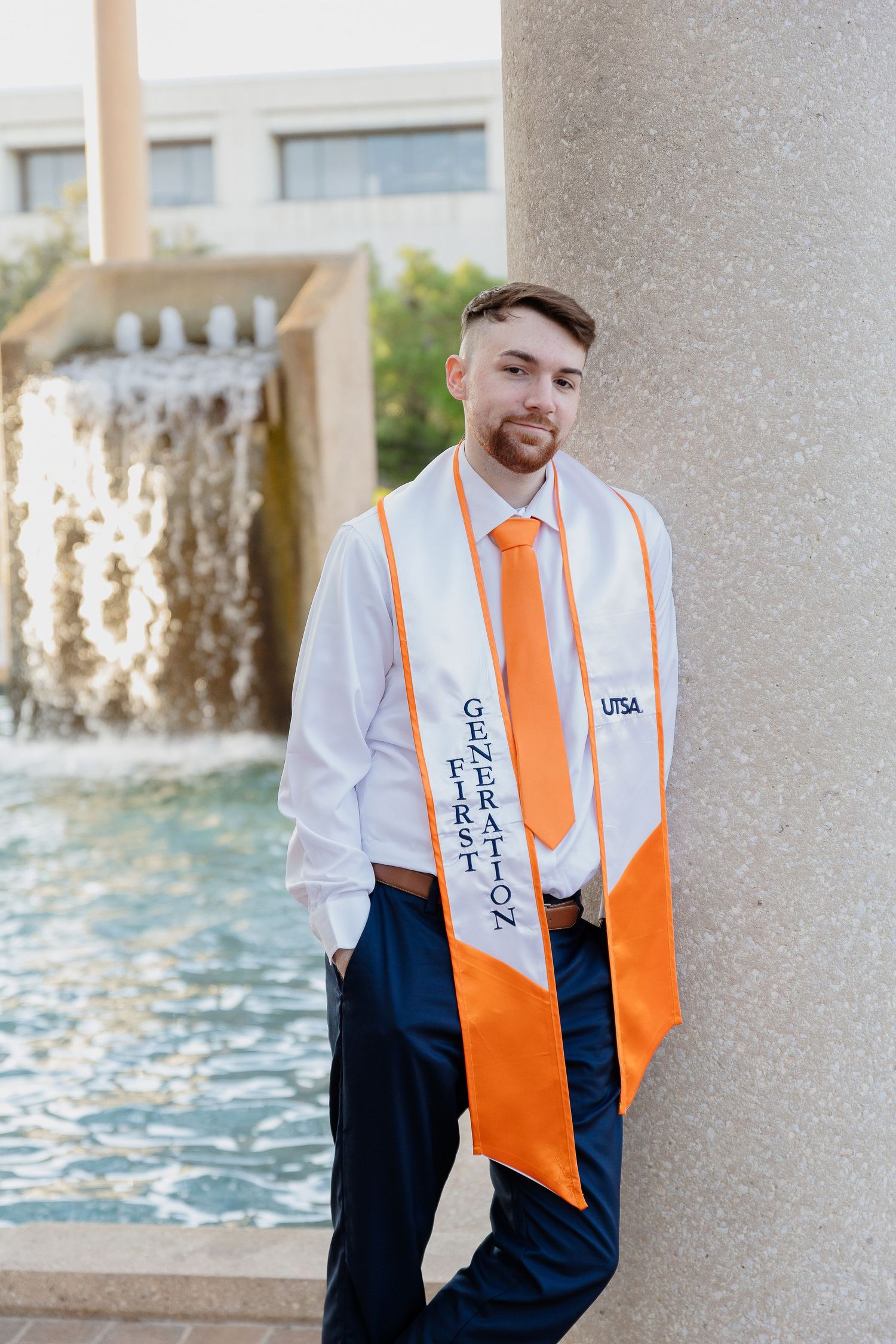 Graduate in formal attire poses by fountain wearing orange graduation stole and tie.