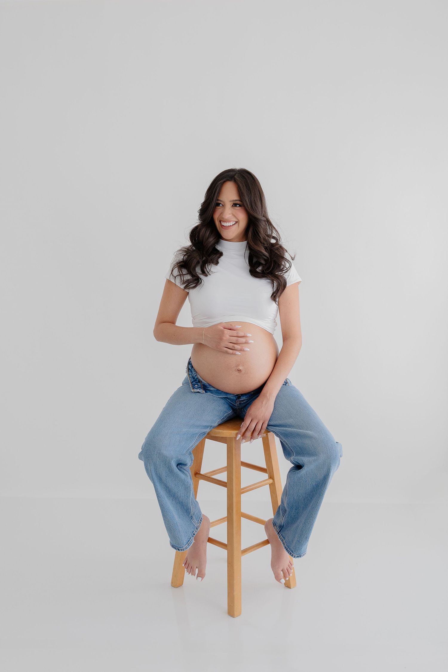 Pregnant woman in white top and blue jeans sits on wooden stool against light gray background in studio setting.