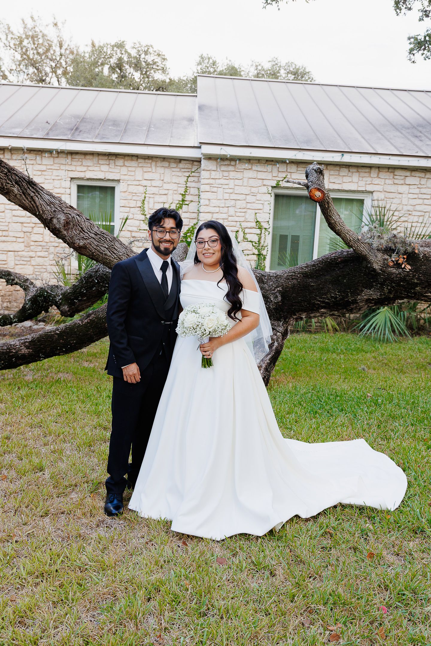 A couple poses together in wedding attire in front of a stone building with green doors and a fallen tree branch.