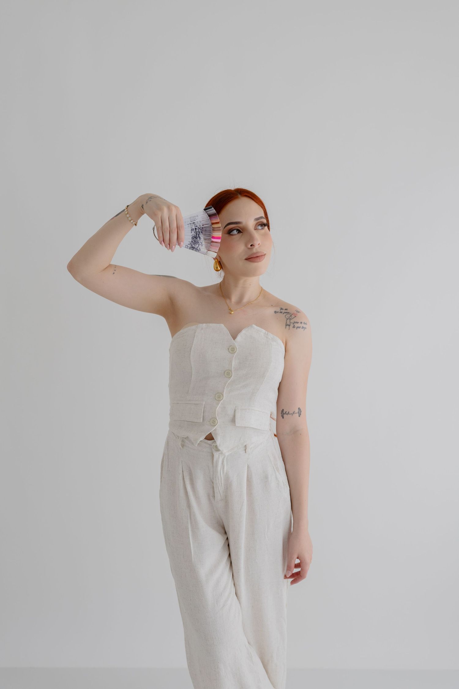 Woman in white strapless jumpsuit poses playfully with vial against light background.
