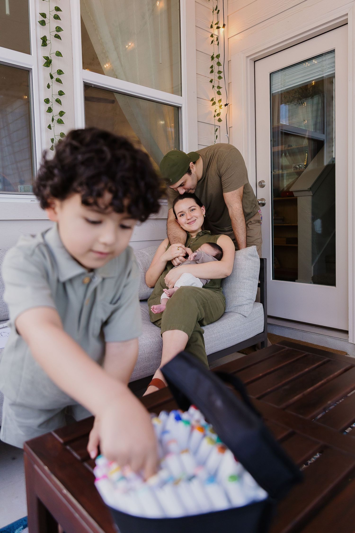 A group sits together on a porch with string lights and vines, while a child interacts with colorful decorations on a table.