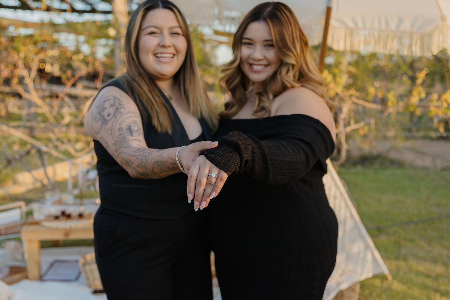 Two friends in black dresses share joyful moment outdoors during sunset.