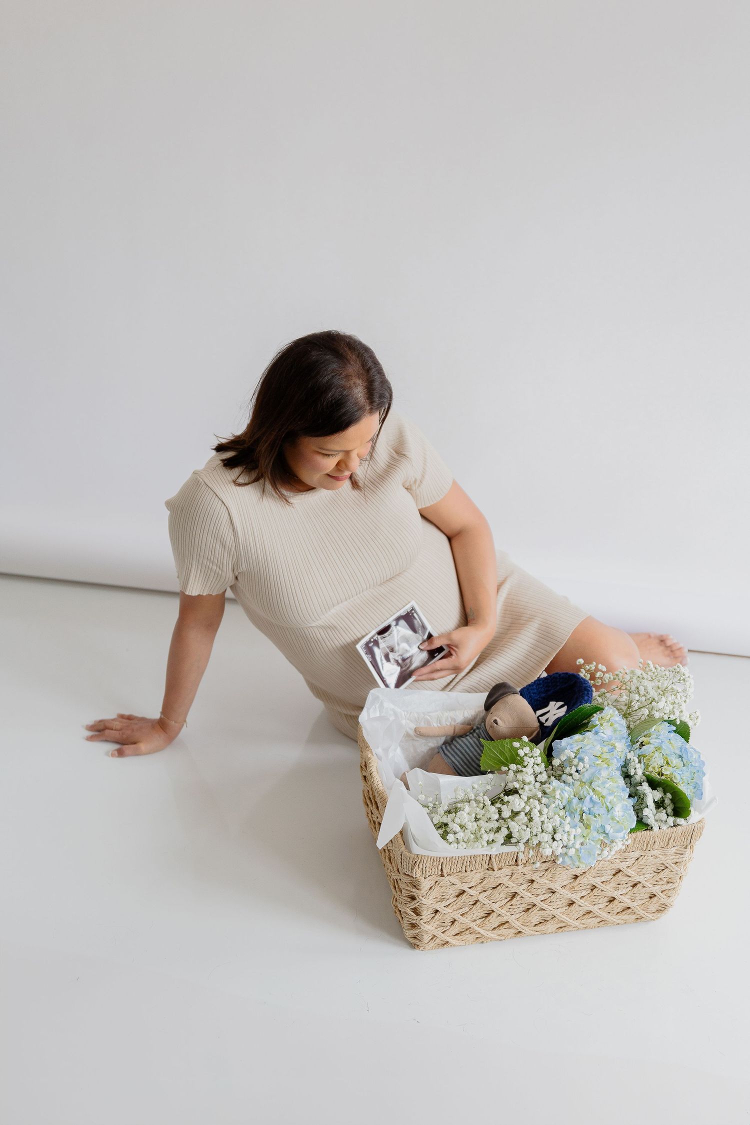 Person in cream dress relaxes beside wicker basket filled with flowers and decor on white floor.