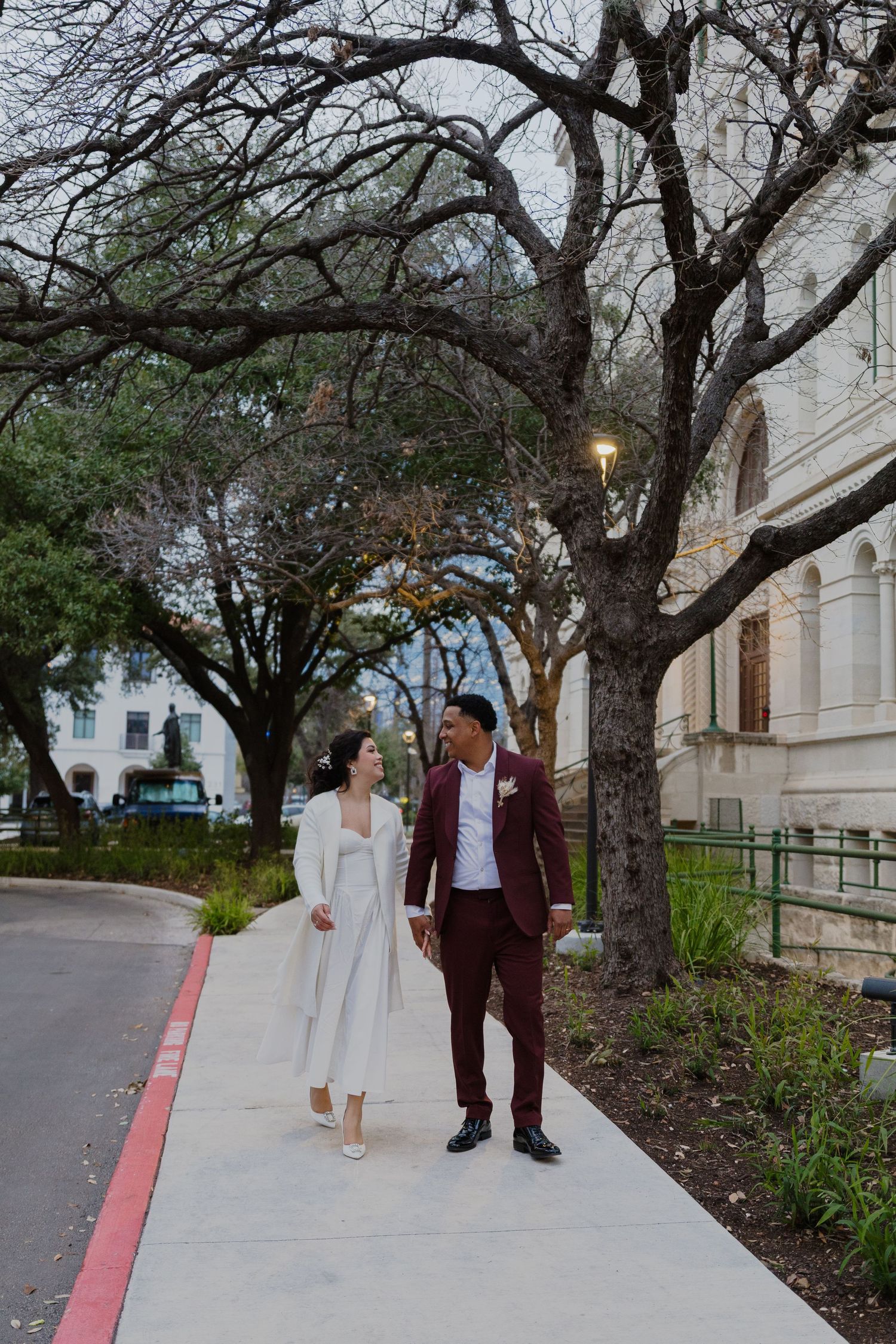 A couple takes romantic walk along tree-lined pathway outside historic building.