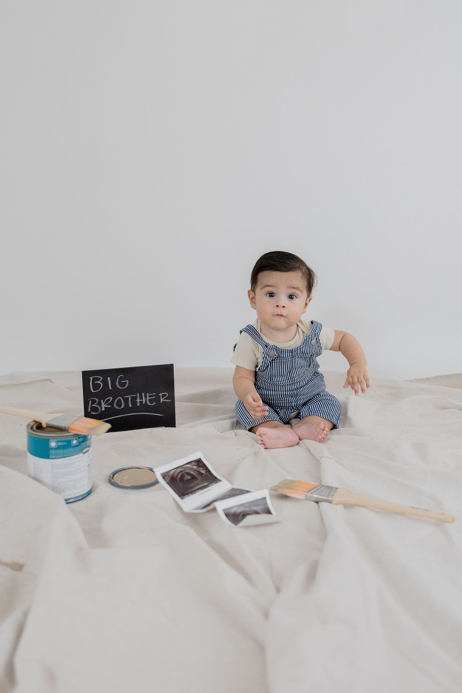 Small child in denim overalls sits on white bedding with ultrasound prints and a chalkboard announcing big brother status.