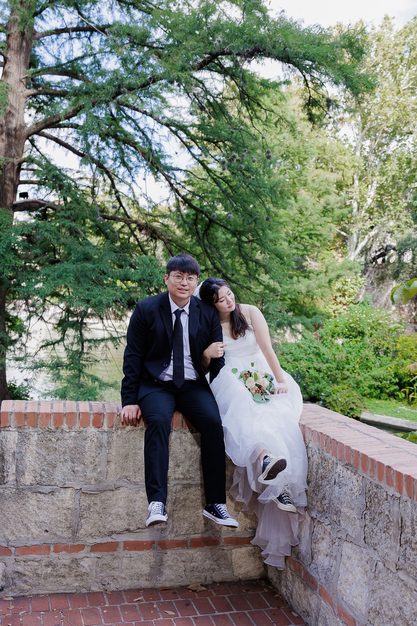 A couple in formal wedding attire sits and poses on a brick wall surrounded by lush green trees.