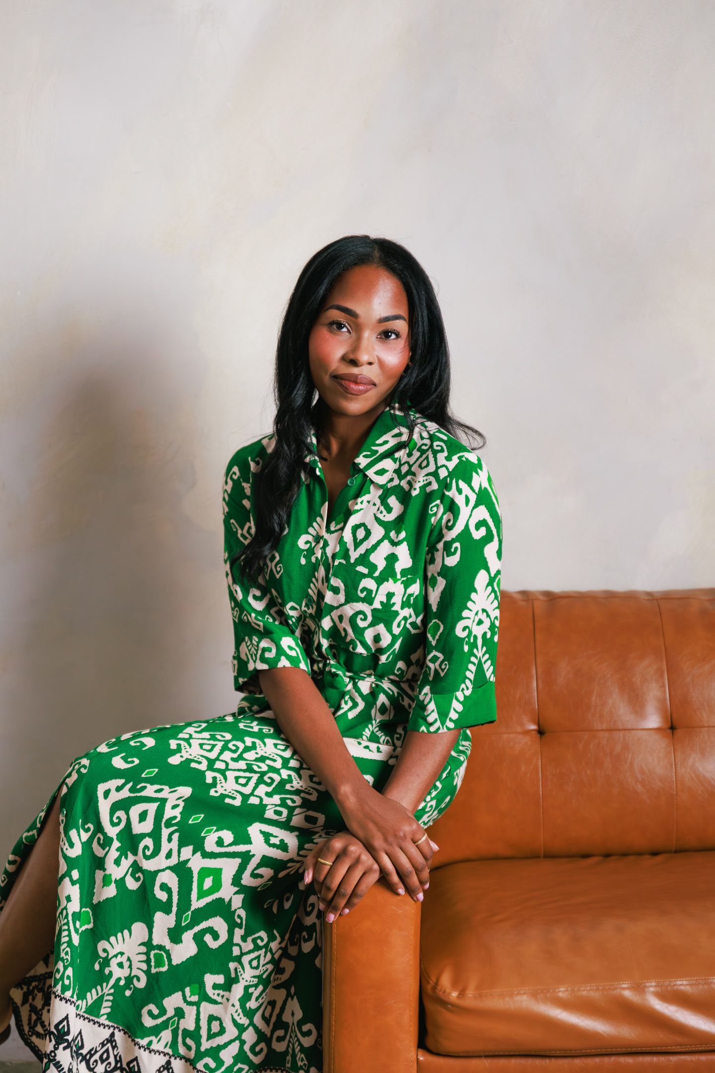 Woman in green and white printed maxi dress seated on a brown leather sofa against a neutral background.