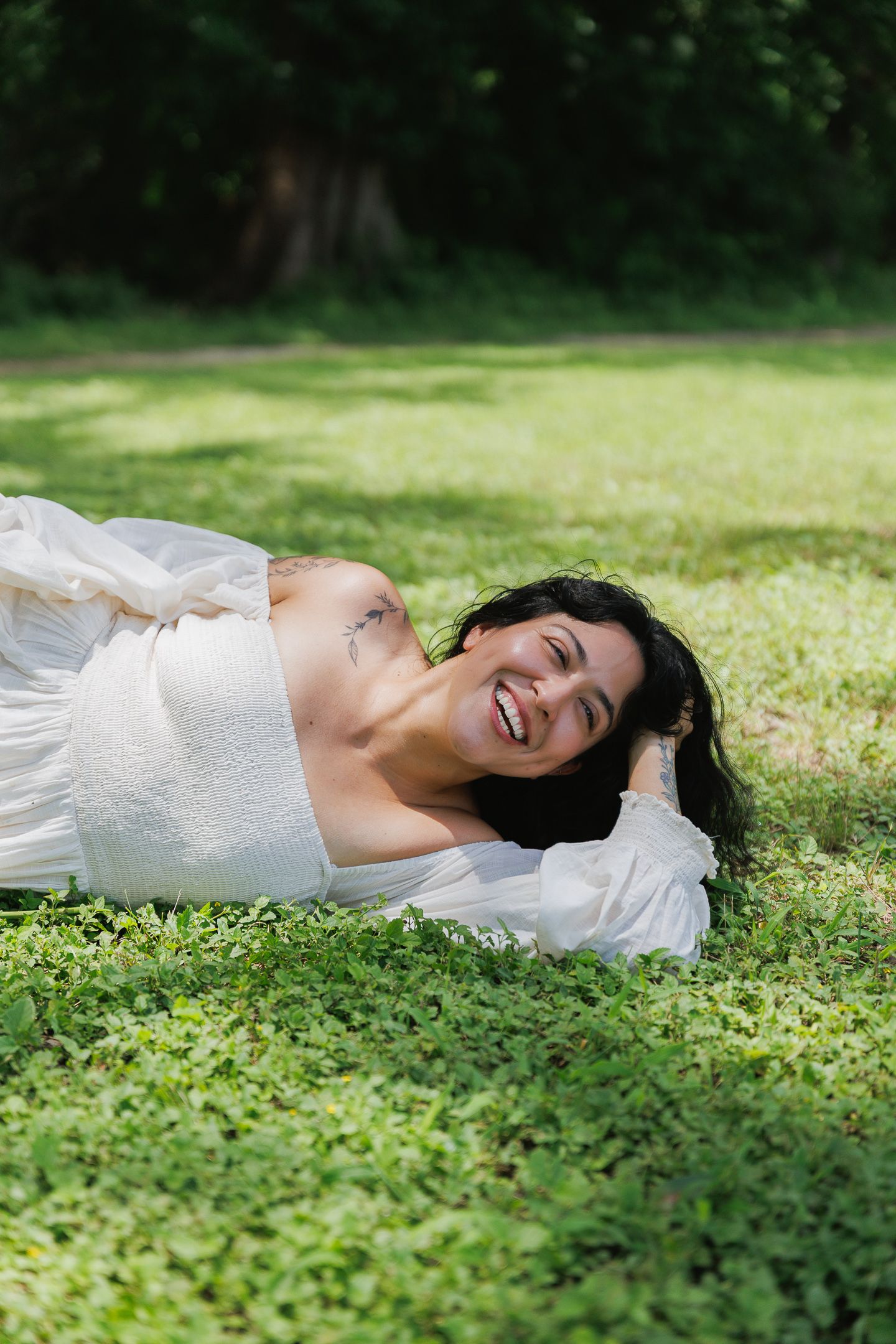 Happy woman in white dress lying on green grass in a sunny park, smiling joyfully at the camera.