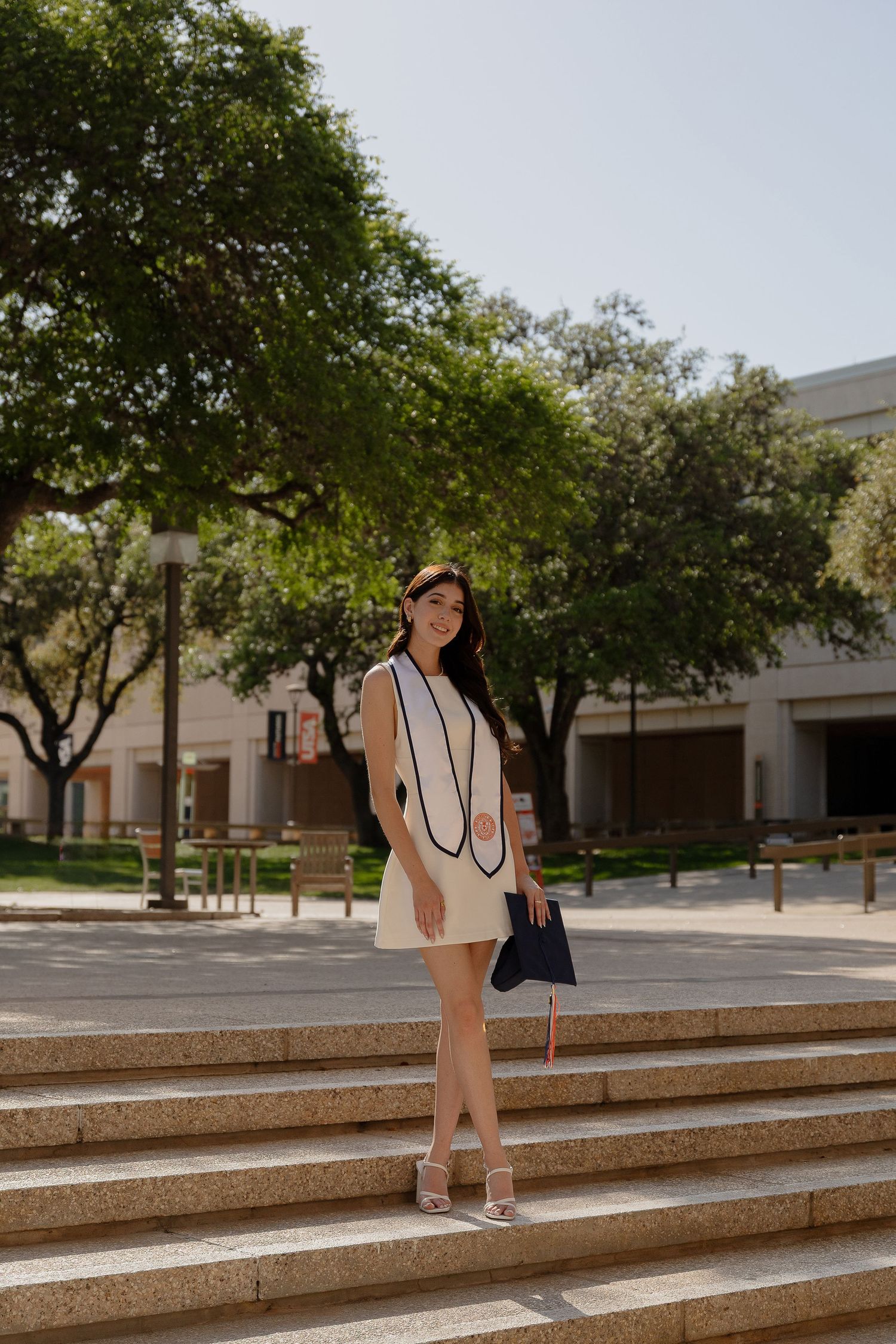 Graduate in cream dress poses on stone steps with trees and campus buildings in background.